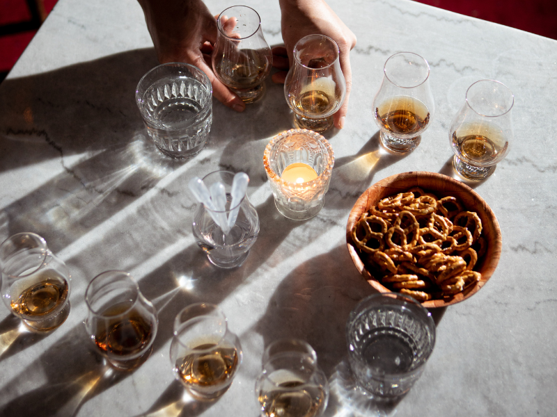 A person’s hand reaching for a glass of alcohol on a table set with multiple glasses of whiskey and water, a bowl of pretzels, and lit candles, with shadows cast on a marble surface.
