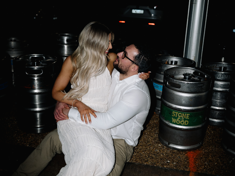 A couple sitting on the ground, embracing and smiling at each other, at a bar or brewery at night with beer kegs in the background.