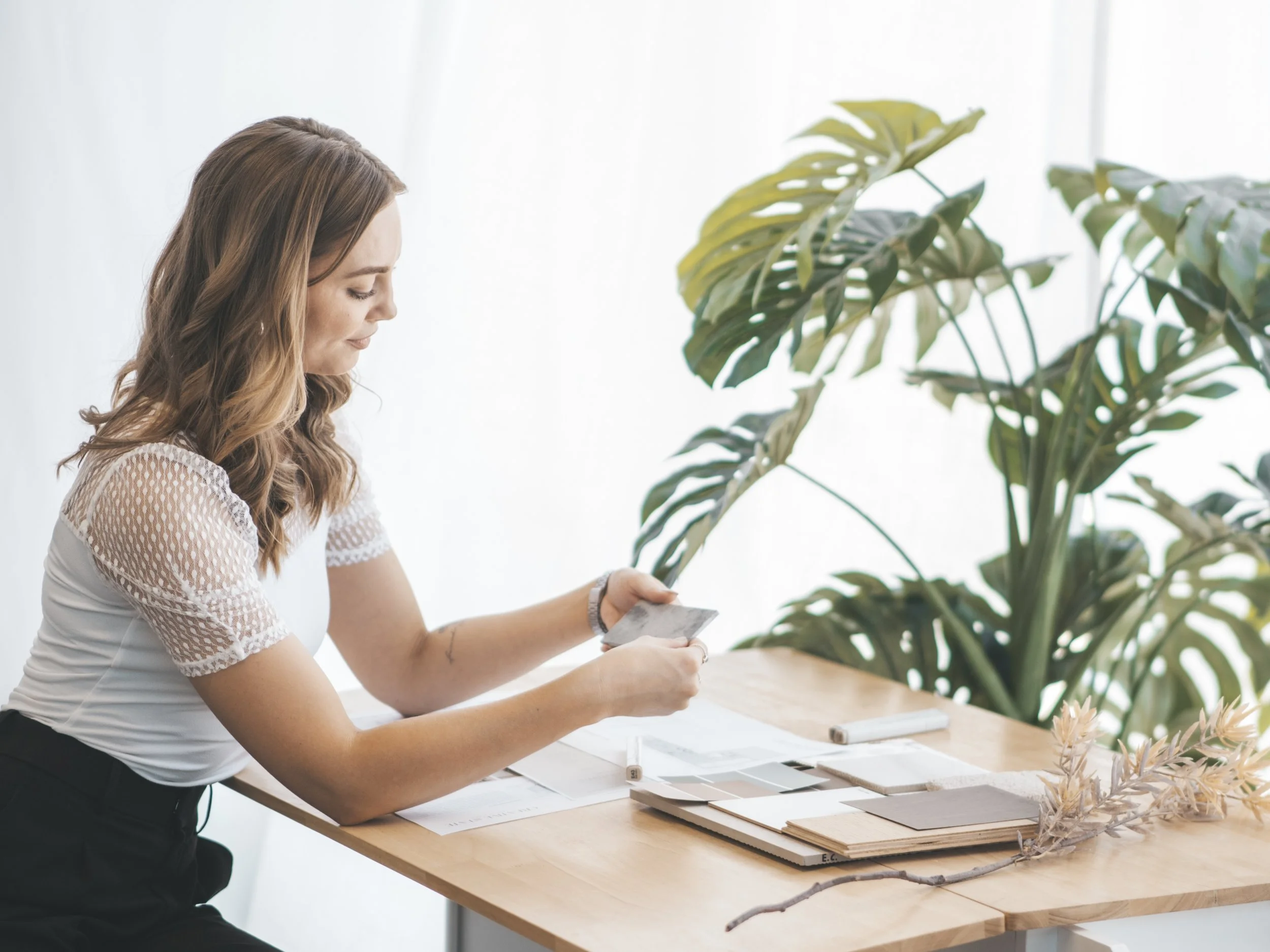 Vanessa, the founder of Creative State Studio, located in Perth, Western Australia is seen sitting at a wooden desk, working with design or color samples, with large green leafy plants in the background.
