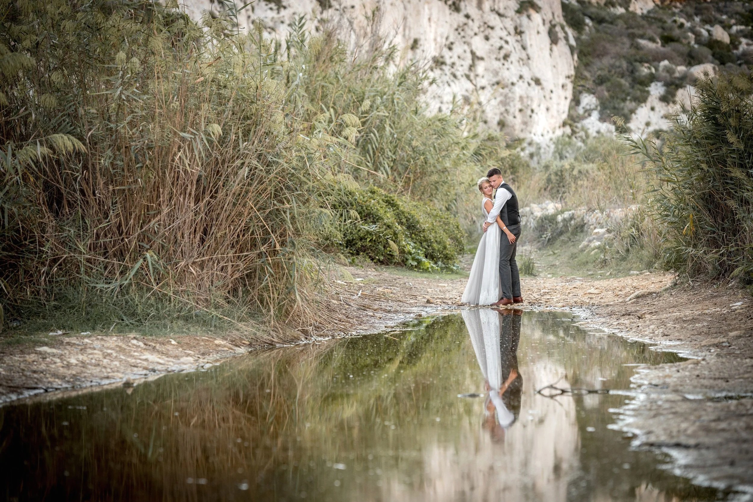 A couple in wedding attire standing close together in a shallow puddle on a dirt path surrounded by tall grasses and rugged cliffs, with their reflection visible in the water.
