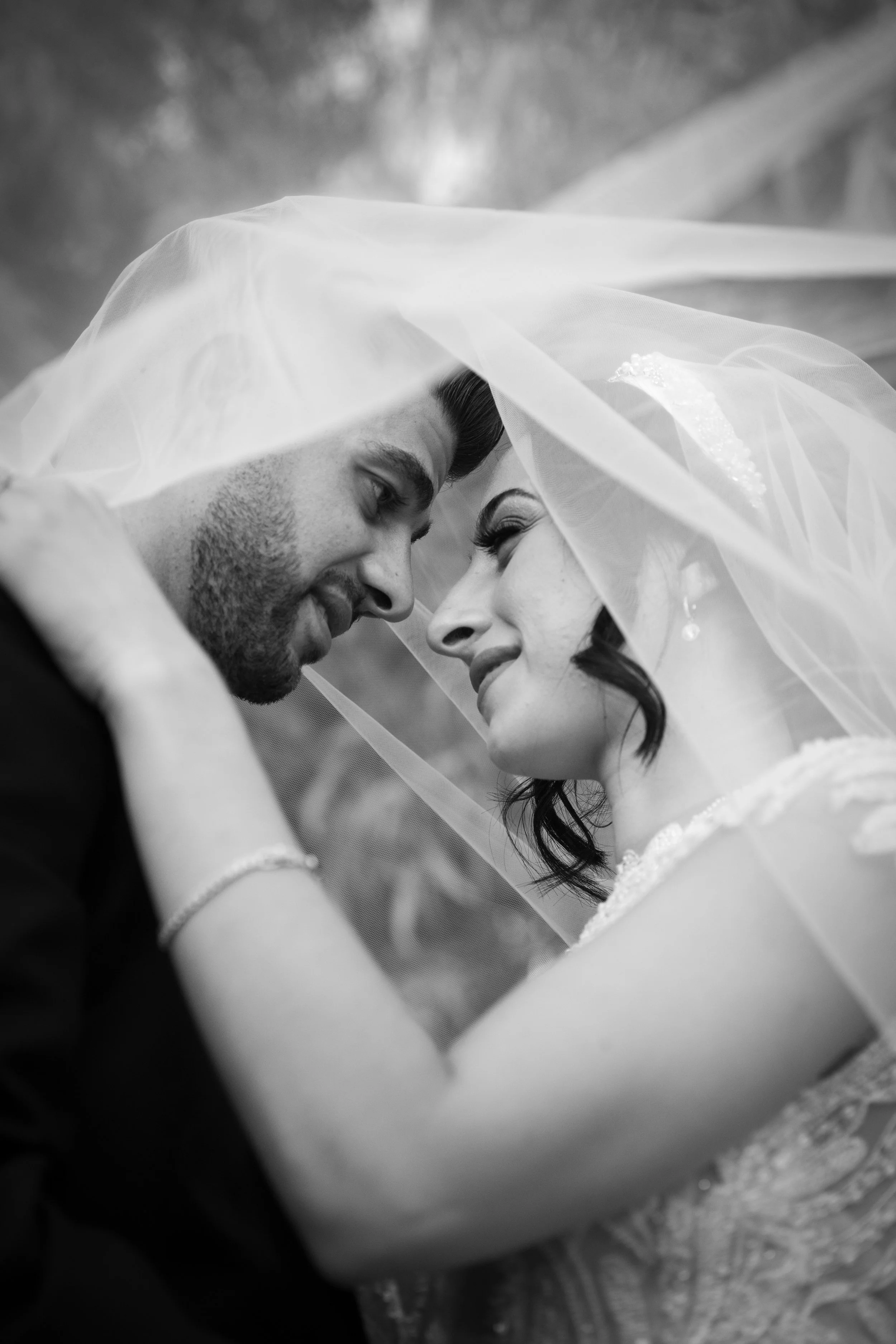 A black-and-white photo of a bride and groom with their foreheads touching, smiling, under a veil, in an intimate moment.