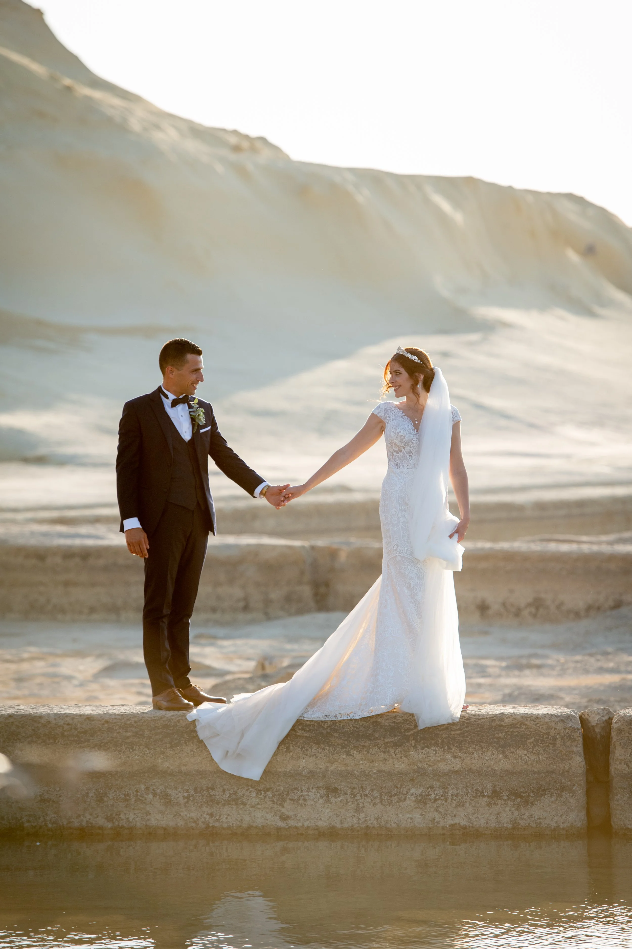 A bride and groom holding hands on a beach during sunset, with large rocks and a ridge in the background.