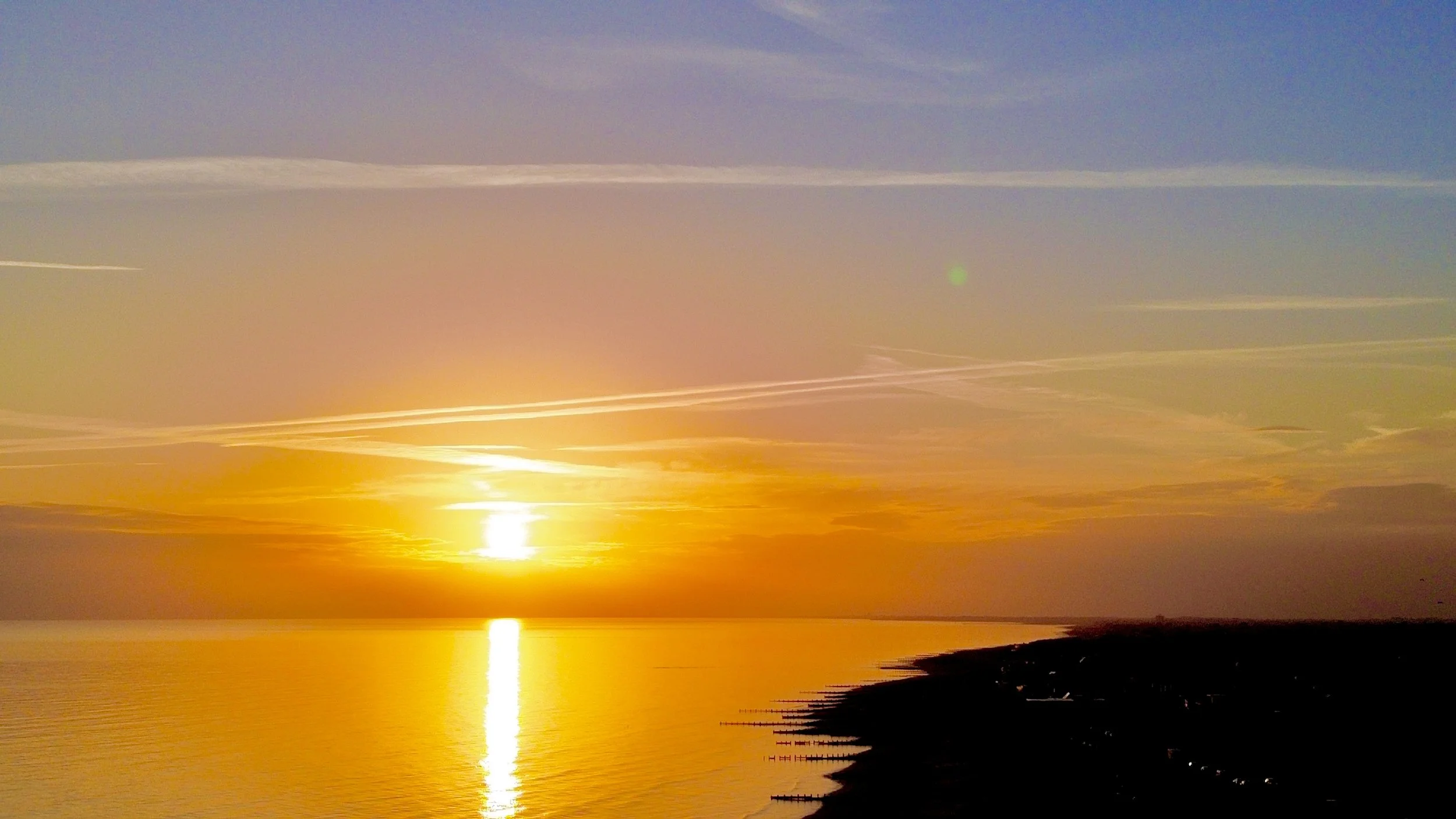 Sunset over calm water with a silhouetted shoreline and some clouds in the sky.