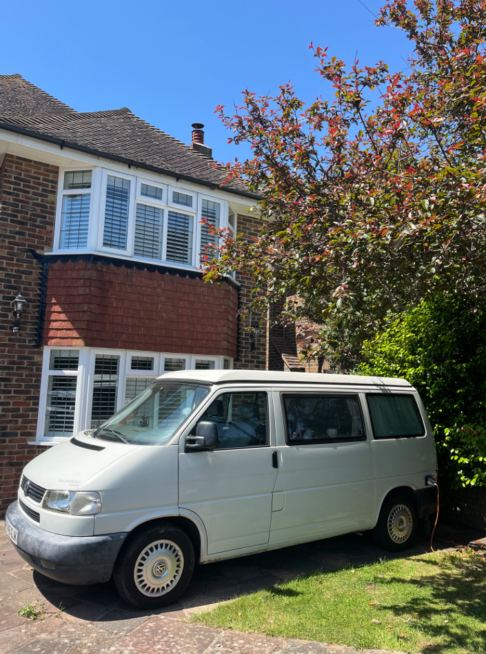 A white van parked in front of a brick house with white window shutters and an overhang. There are trees and bushes nearby under a clear blue sky.