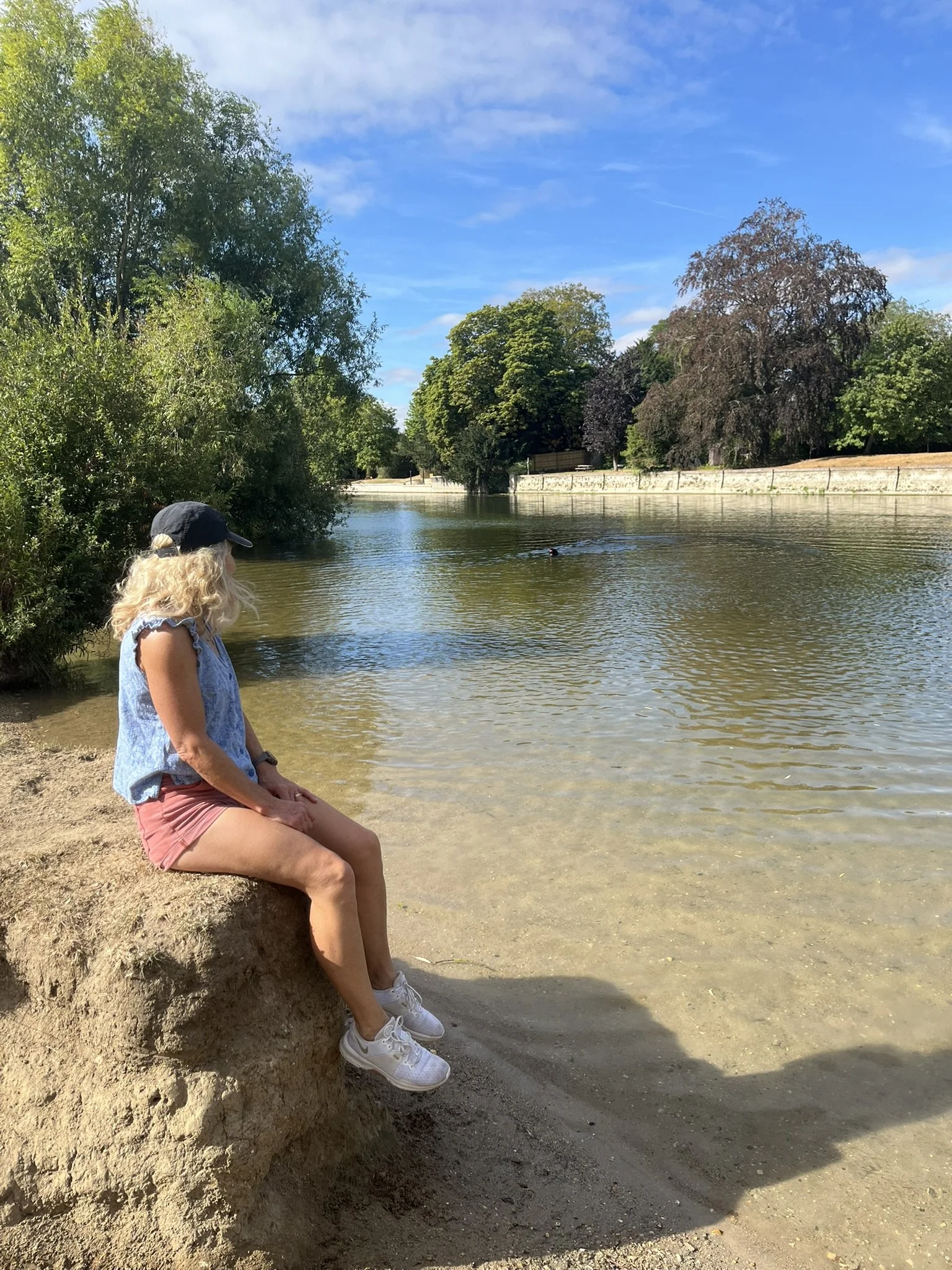 A woman with blond curly hair, wearing a dark baseball cap, a blue sleeveless top, pink shorts, and white sneakers, sitting on a small dirt ledge by a calm river with green trees and a clear blue sky in the background.