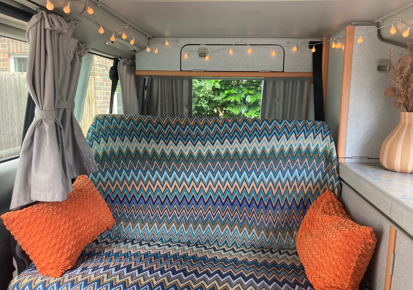 Interior of a camper van with a chevron-patterned blanket on the sofa, orange textured pillows, grey curtains, string lights hanging from the ceiling, and a window with greenery outside.