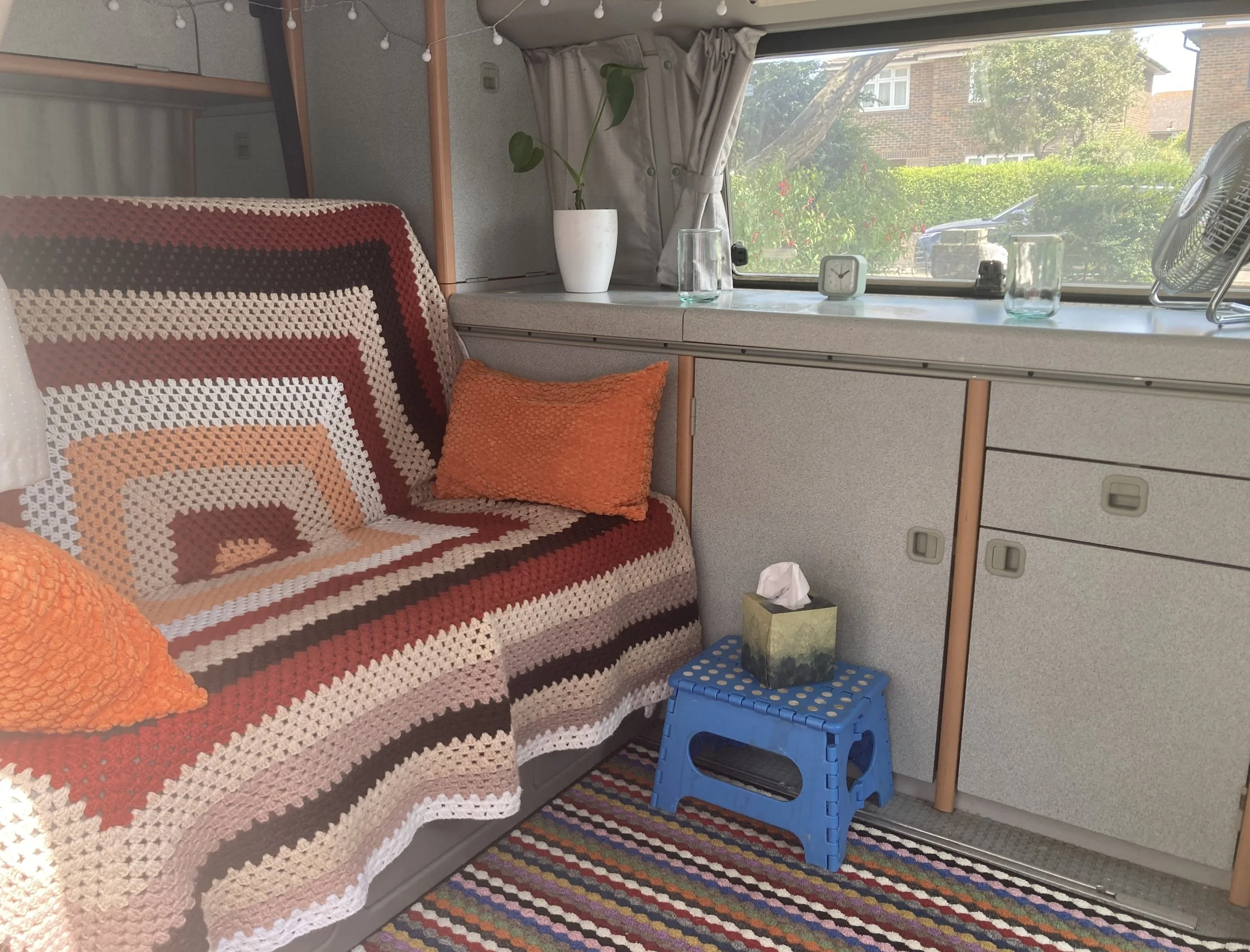 The image shows the interior of a camper or trailer with a window view of houses and greenery outside. Inside, there is a crocheted blanket with a geometric pattern draped over a seating area, orange throw pillows, a small blue step stool, a white va