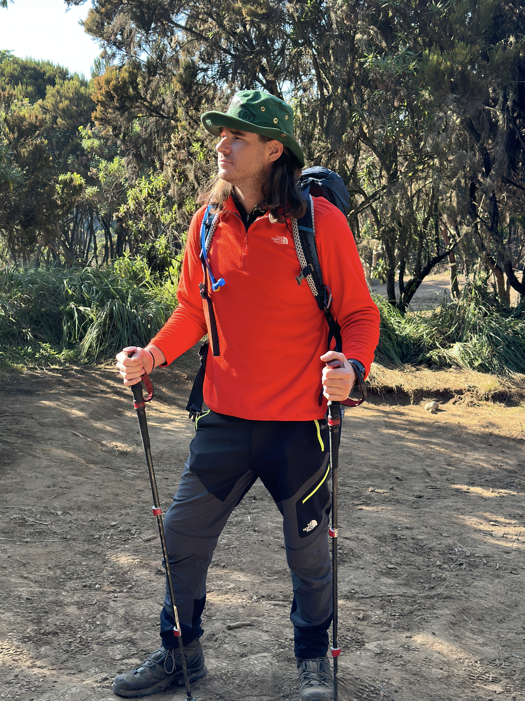 A man with long hair wearing a green hat, red jacket, and hiking gear, including walking poles, standing on a dirt trail in a forested area.