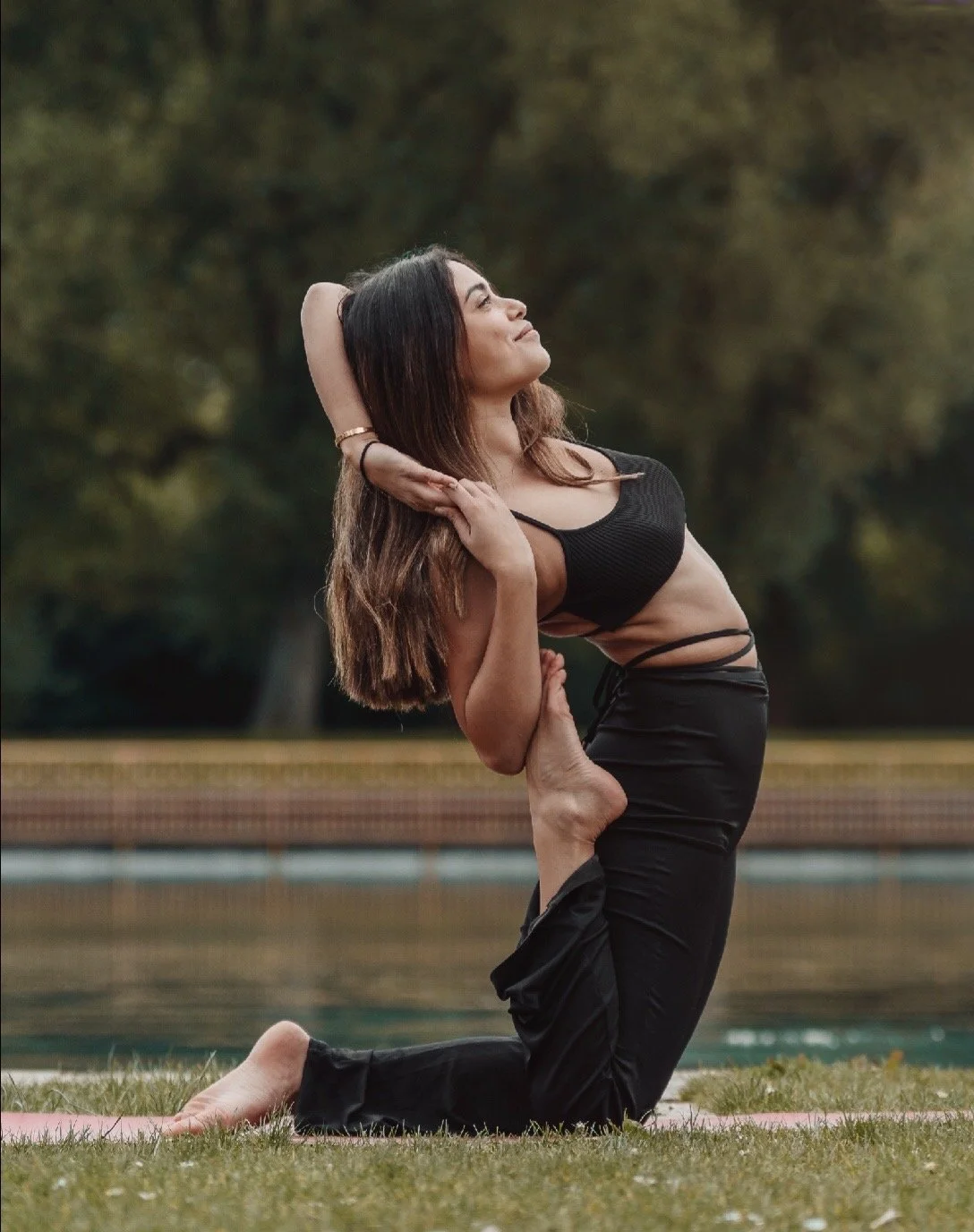 A woman practicing a yoga pose outdoors on a grassy area near a body of water, with trees in the background.