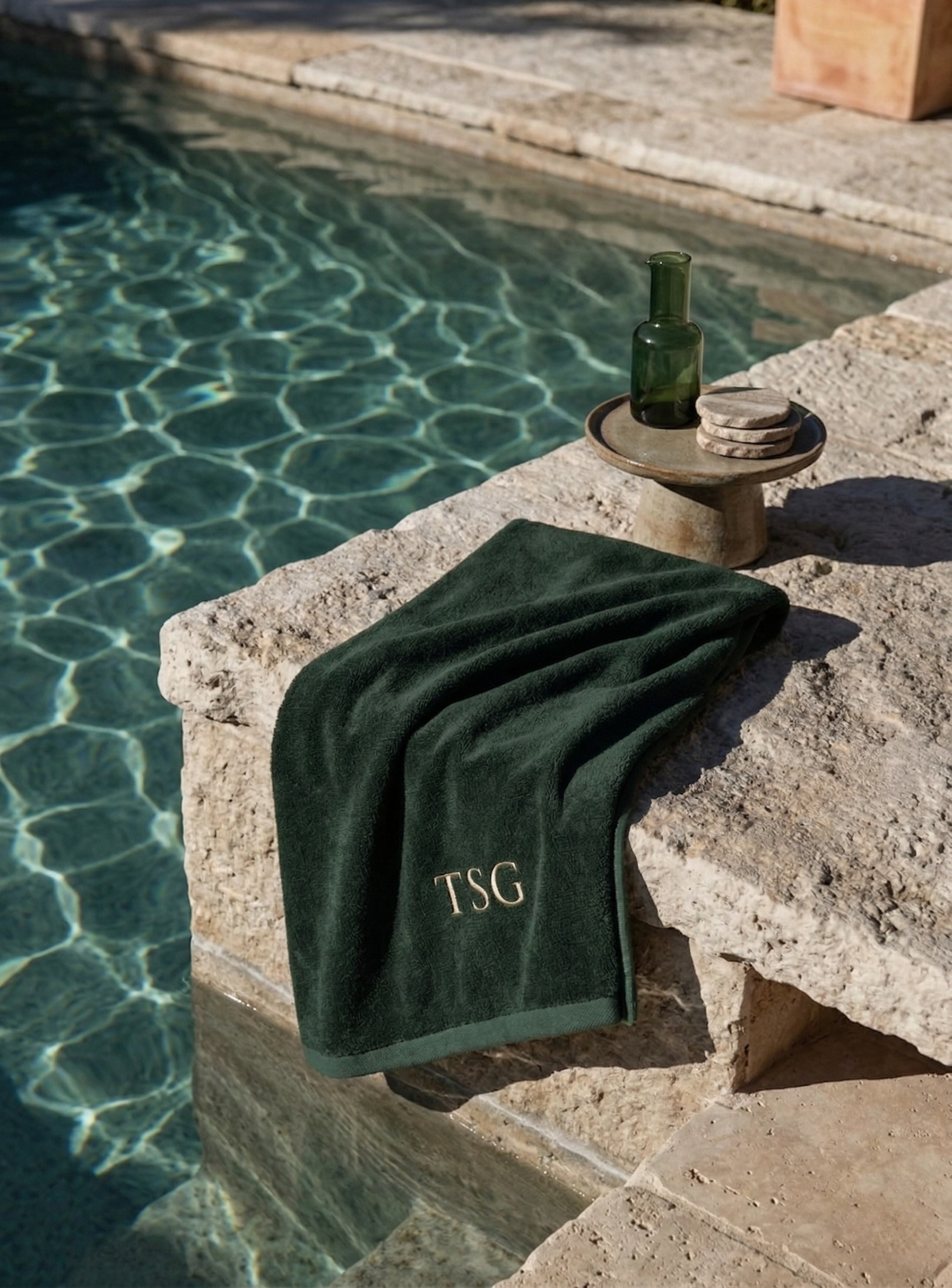 Close-up of a green towel with the initials 'TSG' embroidered on it, draped over a stone poolside ledge. In the background, there is a small round table with a green glass bottle and several stacked stones, beside a swimming pool with clear water reflecting sunlight.
