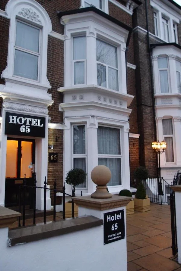 Exterior of Hotel 65, a multi-story brick building with white decorative trim and bay windows, located at 65, with black signs displaying the hotel name and contact information, small potted bushes, a stone railing with a decorative finial, and a lit exterior lamp on the sidewalk.