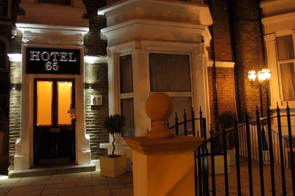 Nighttime exterior view of a hotel building with the sign 'HOTEL 65' illuminated above the entrance. The scene includes brick walls, a black door with orange lighting inside, potted plants, and a decorative street lamp.