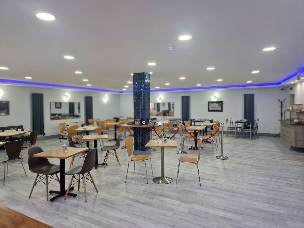 Empty restaurant or dining area with tables and chairs, a blue tiled pillar in the center, and blue lighting along the ceiling.