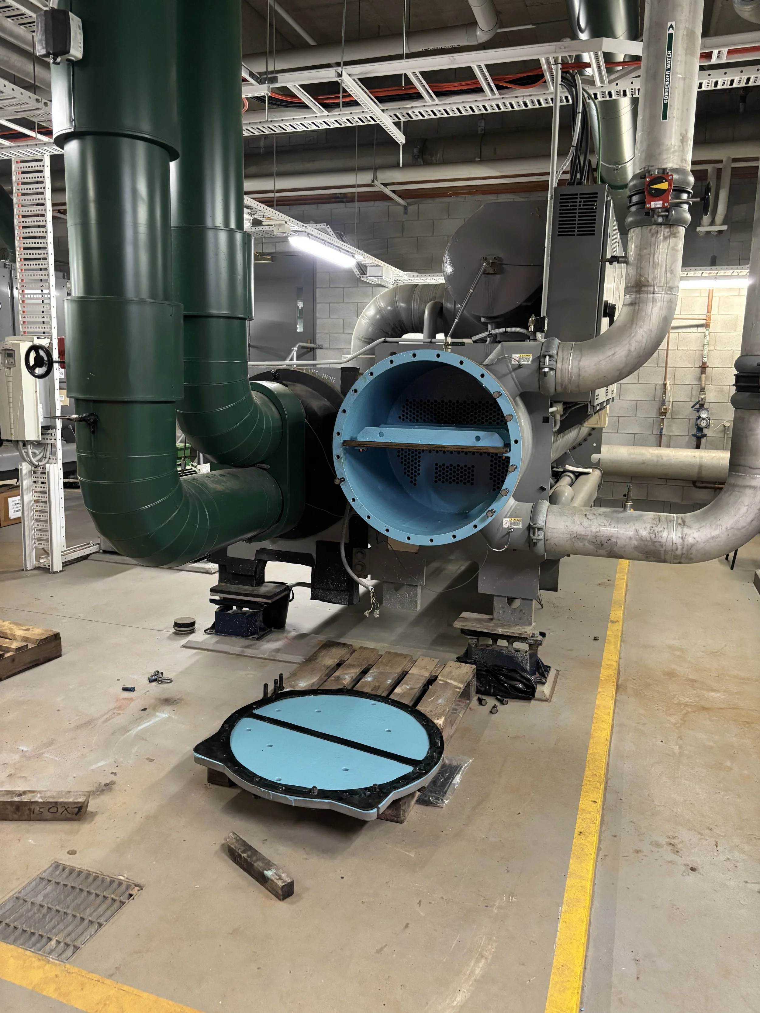 Industrial machinery with large green and silver pipes in a factory setting, with a blue access hatch and various tools and wooden pallets on the floor.