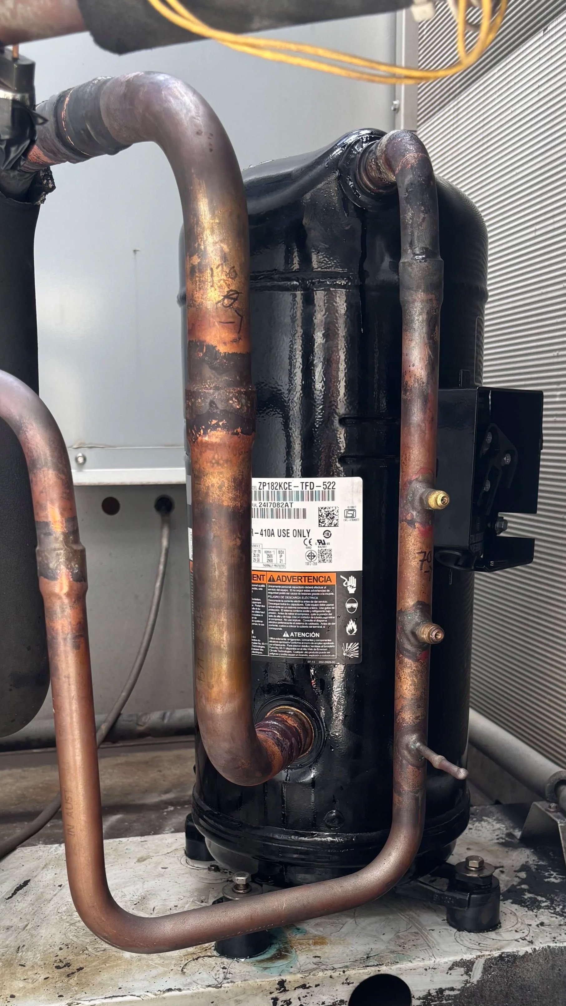 Close-up of a black refrigeration compressor with copper pipes, some showing signs of rust and corrosion, mounted on a white surface with a gray background.