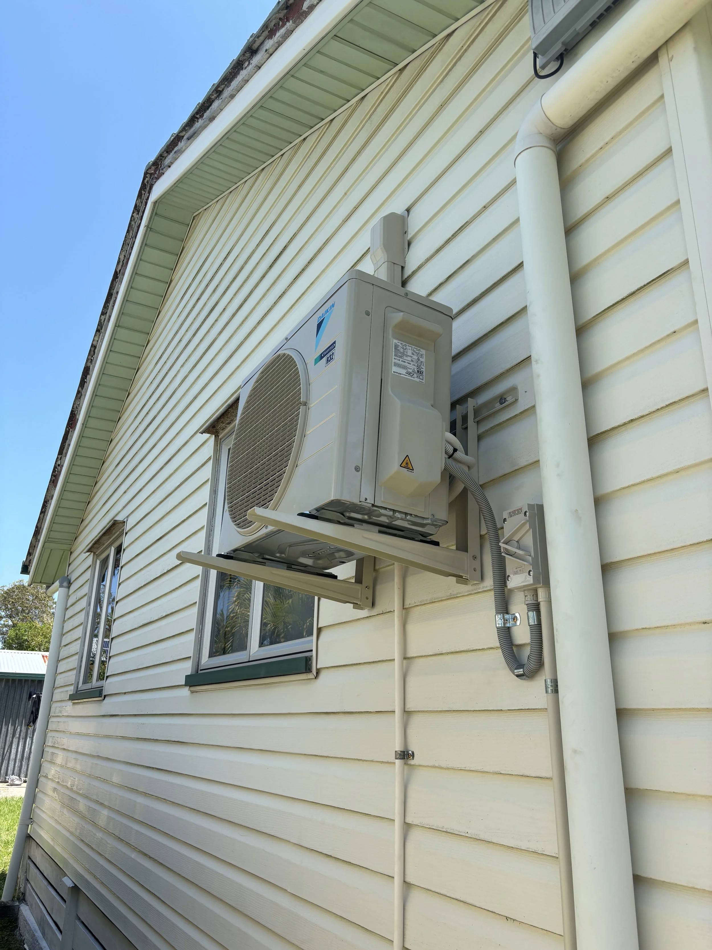 Air conditioning unit installed on the exterior wall of a house with vinyl siding, two windows, and a gutter pipe, under a clear blue sky.