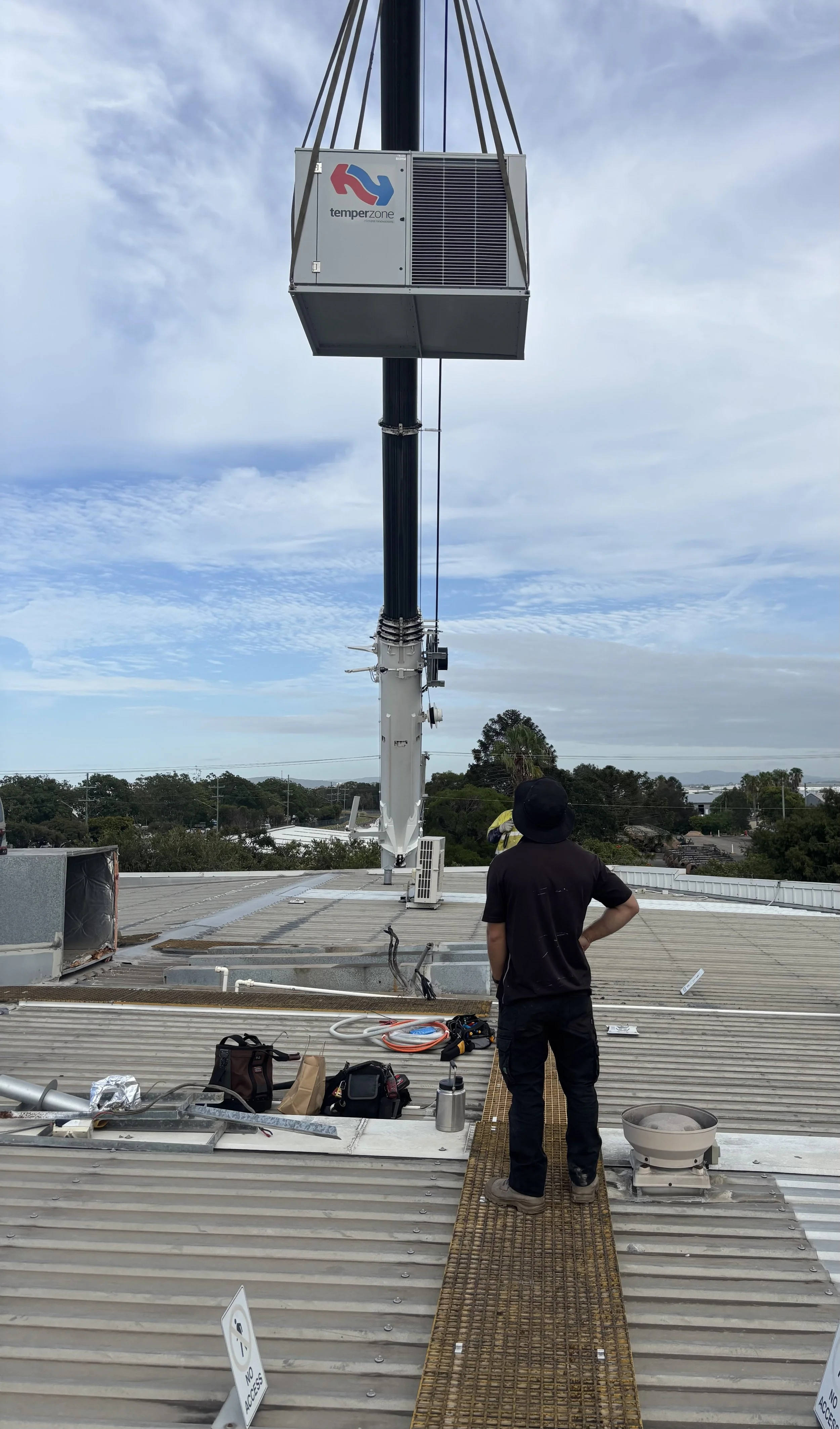 A person in black clothing and a hat stands on a metal walkway on a rooftop, observing an HVAC unit being installed or maintained on a tall antenna mast. Tools and equipment are scattered on the roof.