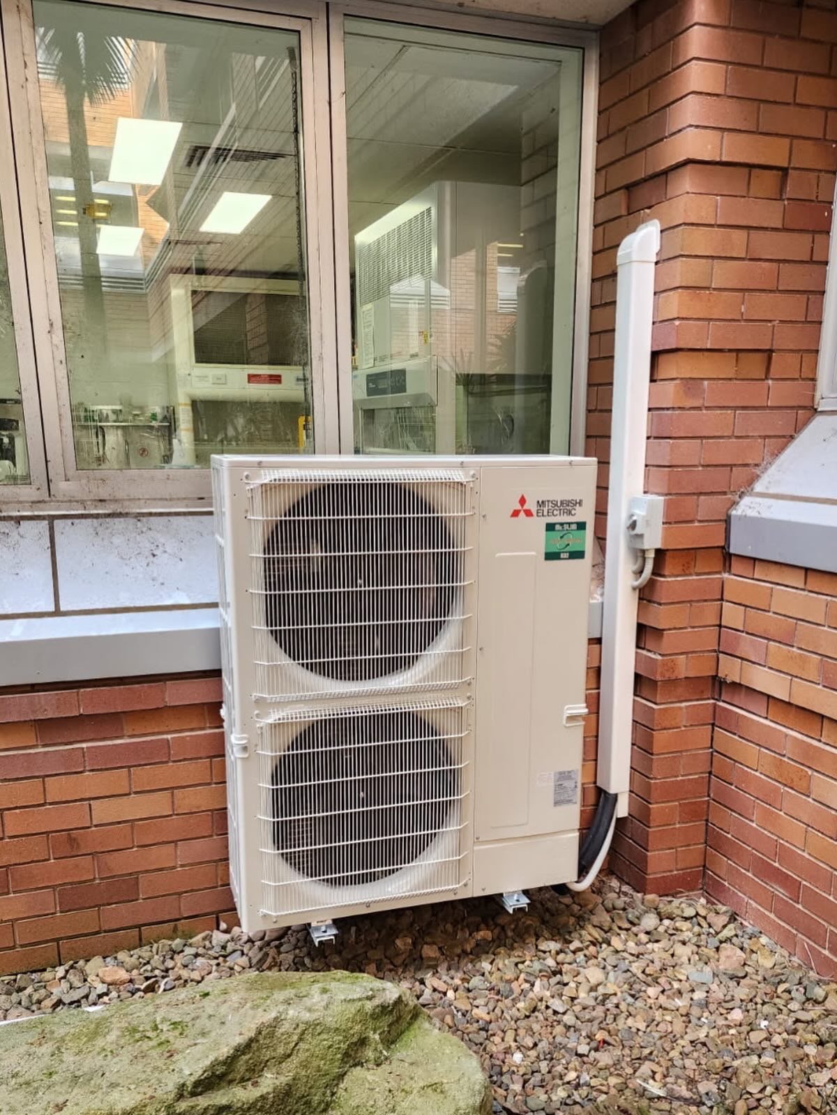Outdoor Mitsubishi Electric air conditioning unit mounted on the brick exterior wall of a building, with rocks on the ground below.