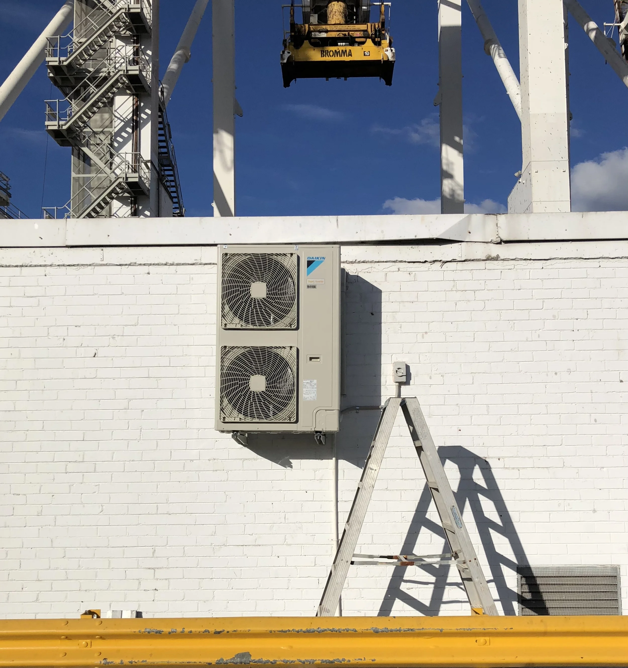 A white brick wall with an air conditioning unit mounted on it, a ladder leaning against the wall, and a yellow barrier at the bottom. In the background are structural elements of a Ferris wheel and a clear blue sky.