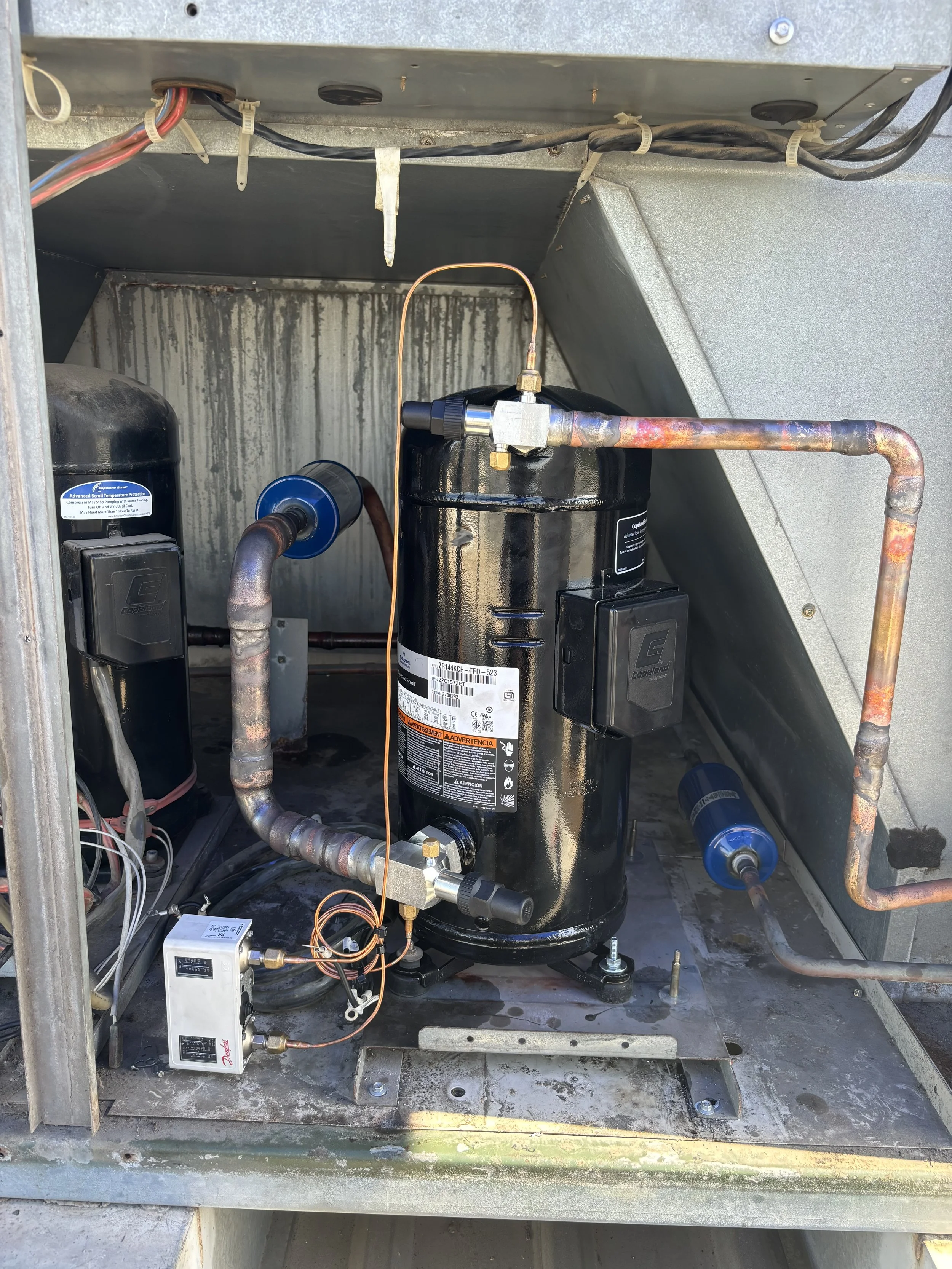 A close-up view of an HVAC compressor unit with copper piping, electrical wiring, and components inside an outdoor metal enclosure.
