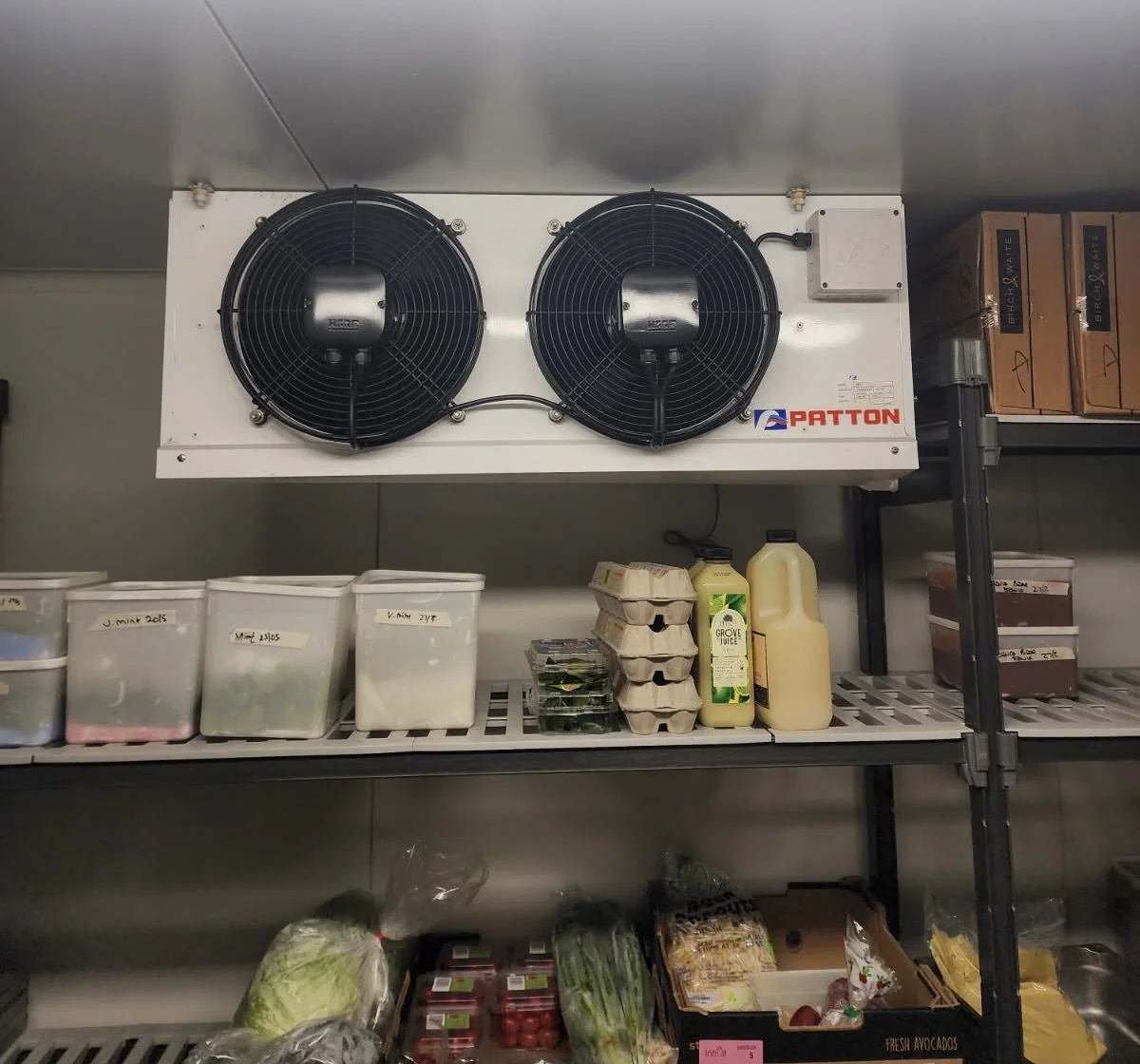 Industrial refrigerator fan unit with two large black fans and a PATTON logo, second shelf with storage containers, eggs, juice, and vegetables, bottom shelf with packaged produce and vegetables.