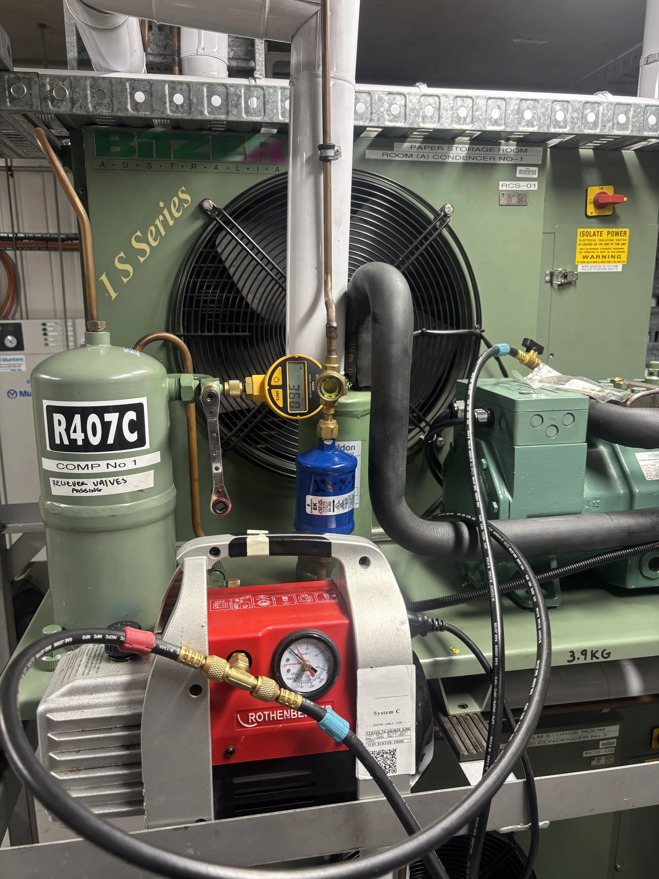 Industrial refrigeration system with various components including a green compressor, a red Rothenberger vacuum pump, gauges, hoses, and electrical wiring, set in a mechanical room.