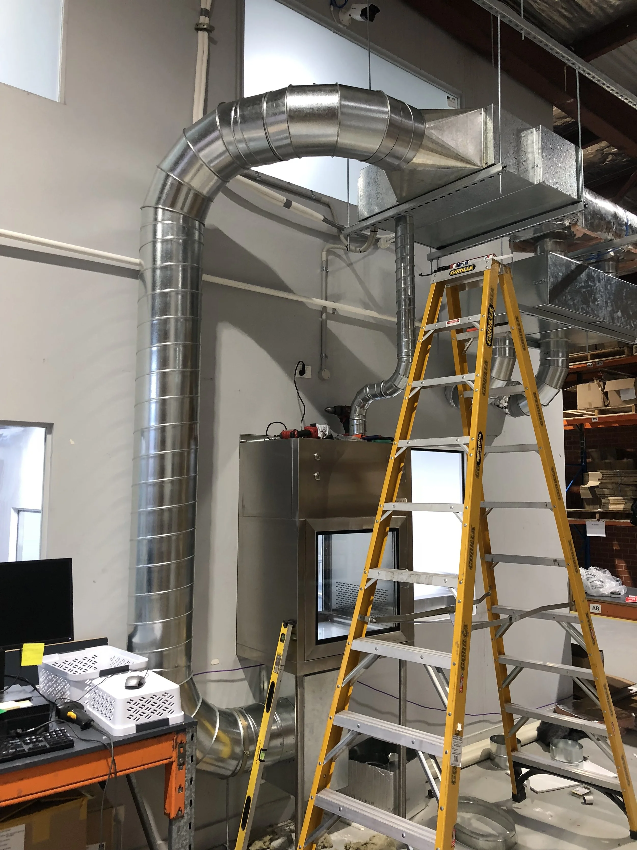 Industrial ductwork and ventilation system installation with a tall yellow ladder in an office or workshop setting.