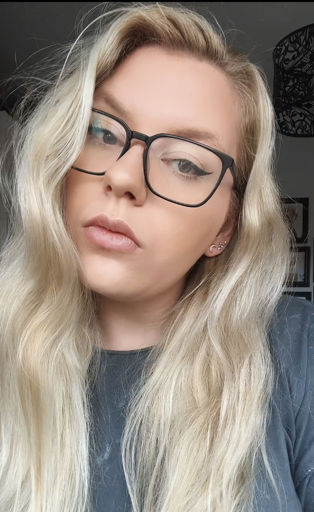 A young woman with long, wavy blonde hair and black-rimmed glasses looking into the camera.