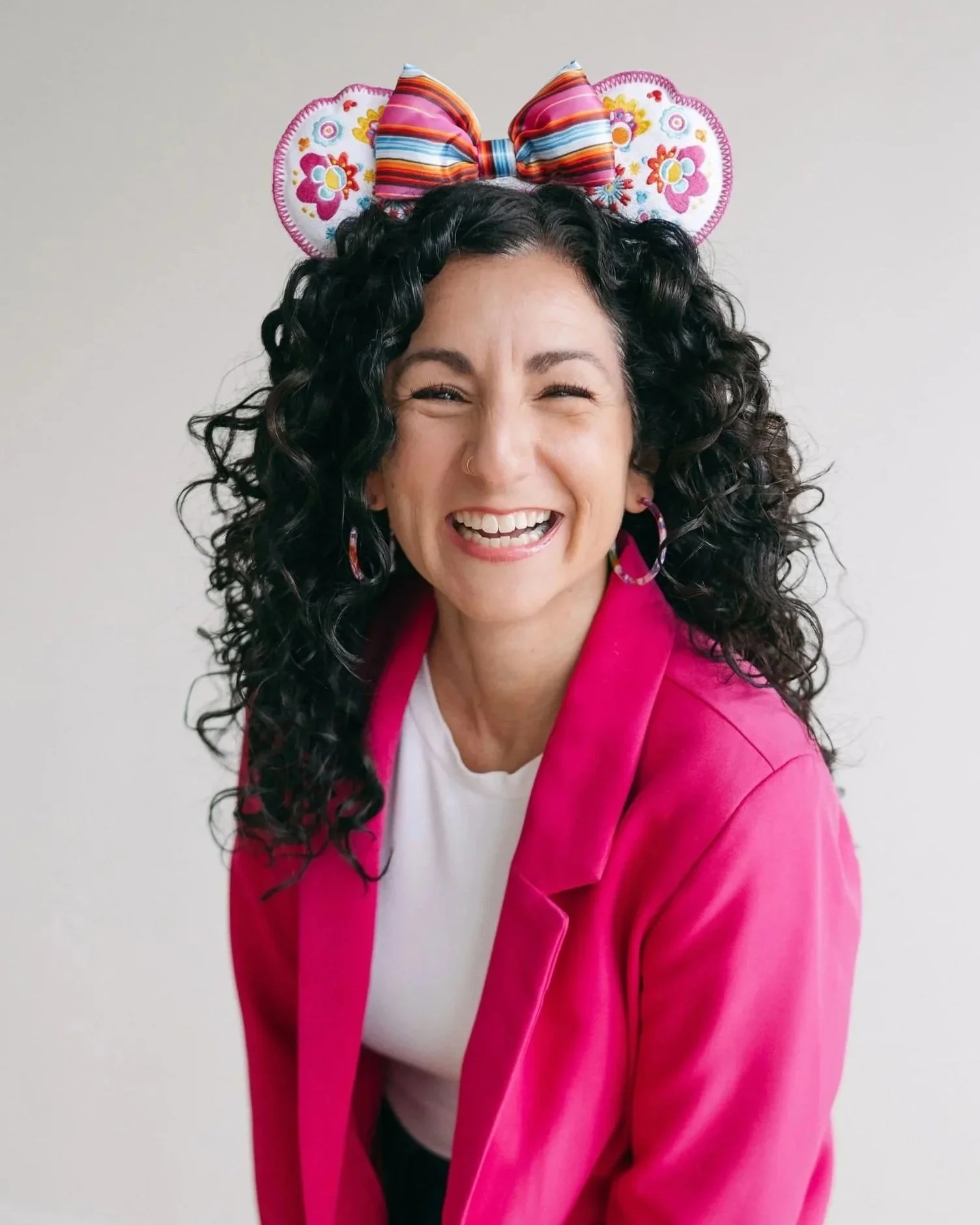A woman with black curly hair, wearing a pink blazer, white shirt, smiling, with colorful striped bow headband with floral embroidered ears.
