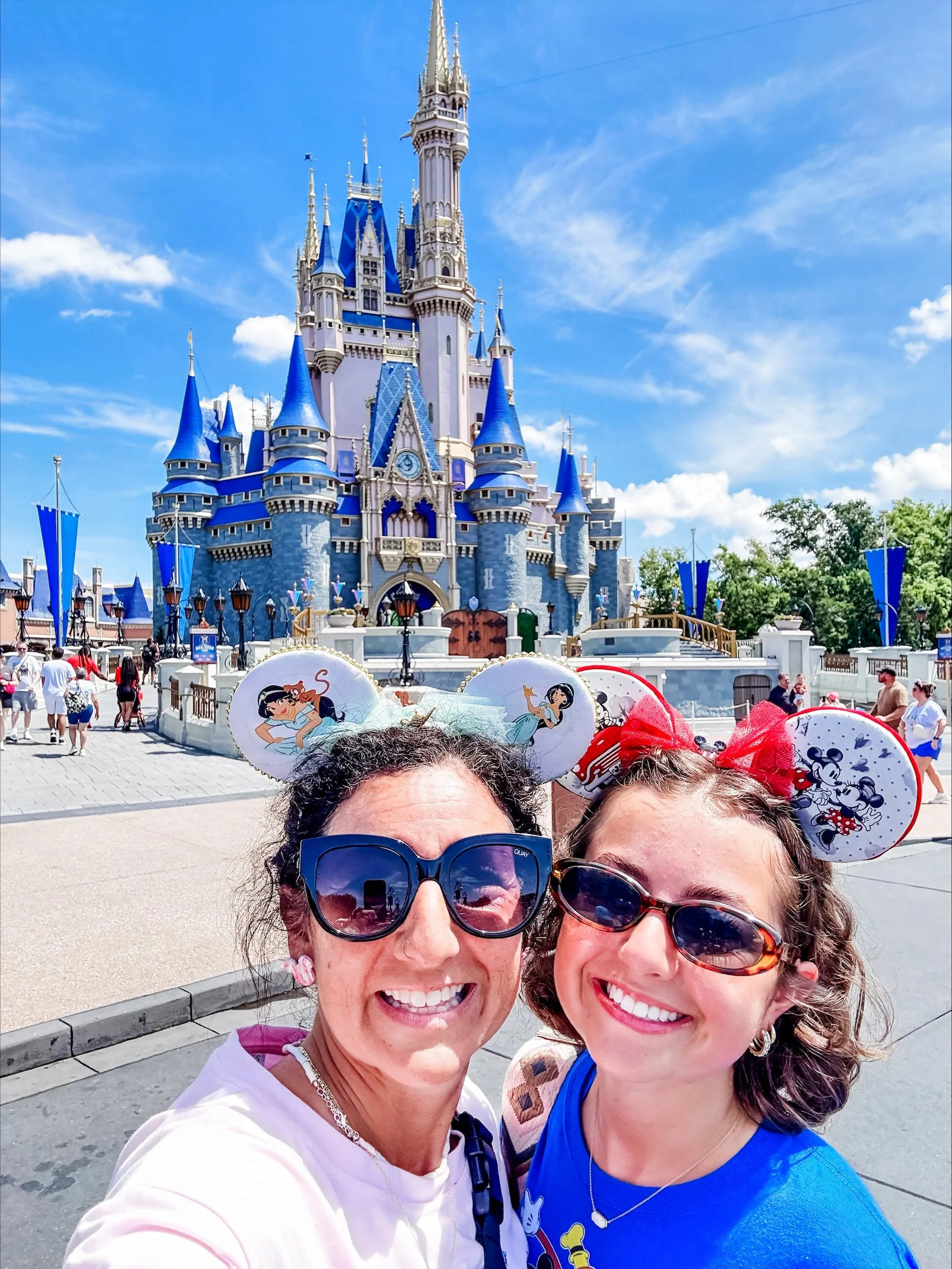 Two women smiling for a selfie in front of the Cinderella Castle at a Disney theme park, wearing Mickey Mouse ear hats and sunglasses.
