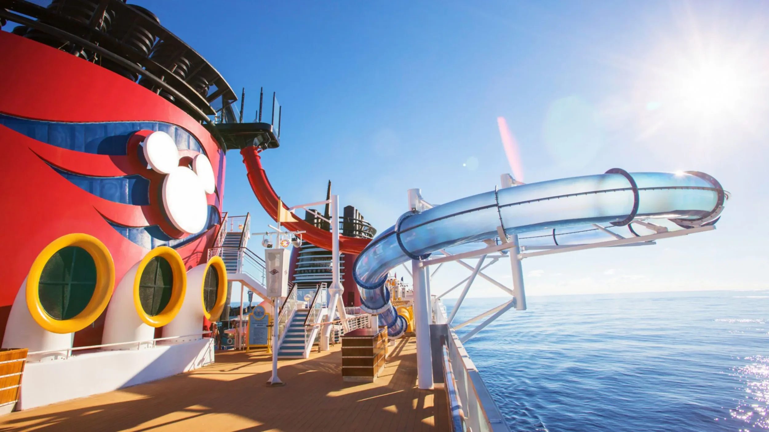 View of a cruise ship deck with a giant Mickey Mouse-themed water slide and water attractions, overlooking the ocean under a bright, sunny sky.