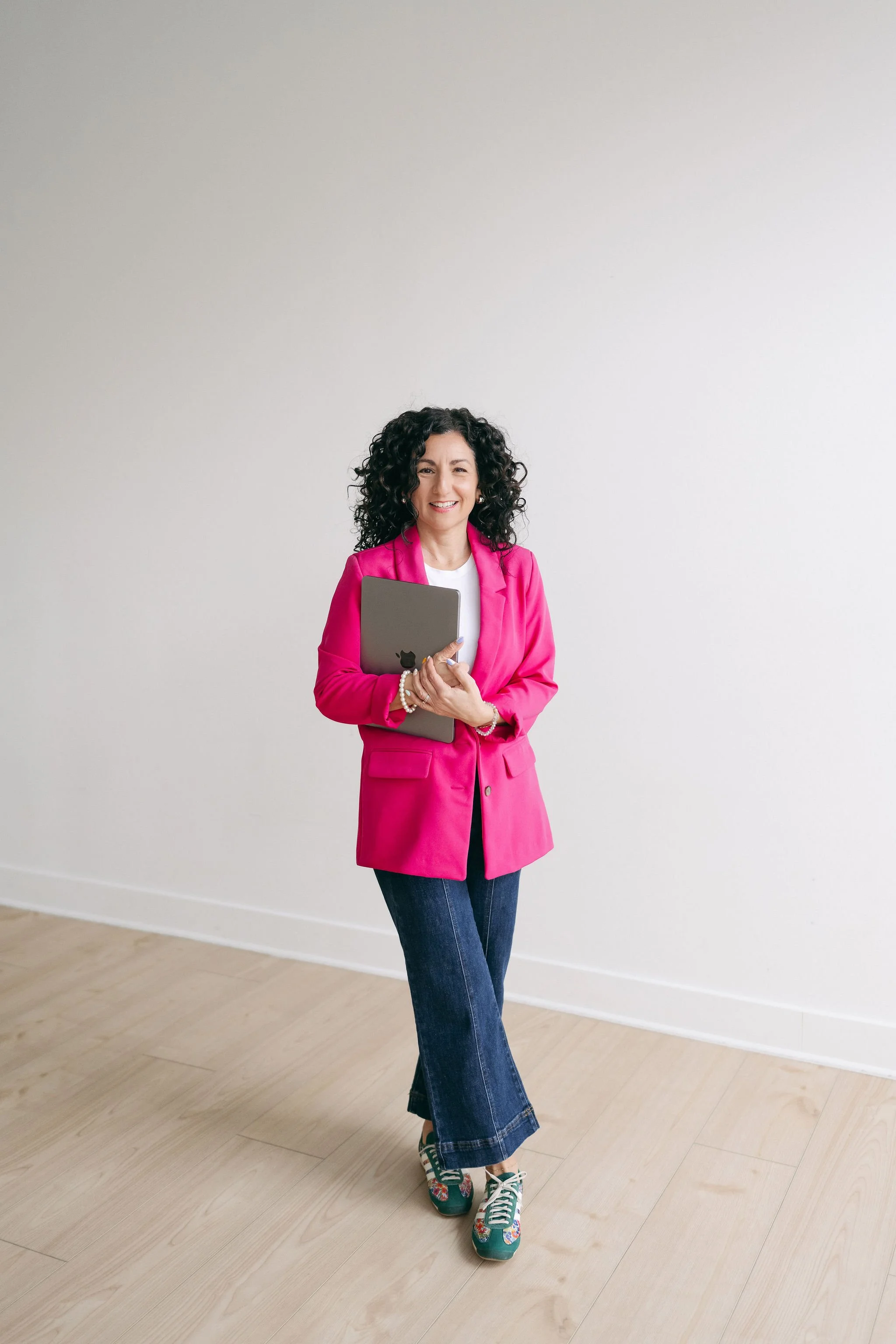 A woman with curly dark hair, wearing a bright pink blazer, white shirt, dark wide-leg jeans, and colorful sneakers, standing in front of a plain white wall and holding a silver laptop.