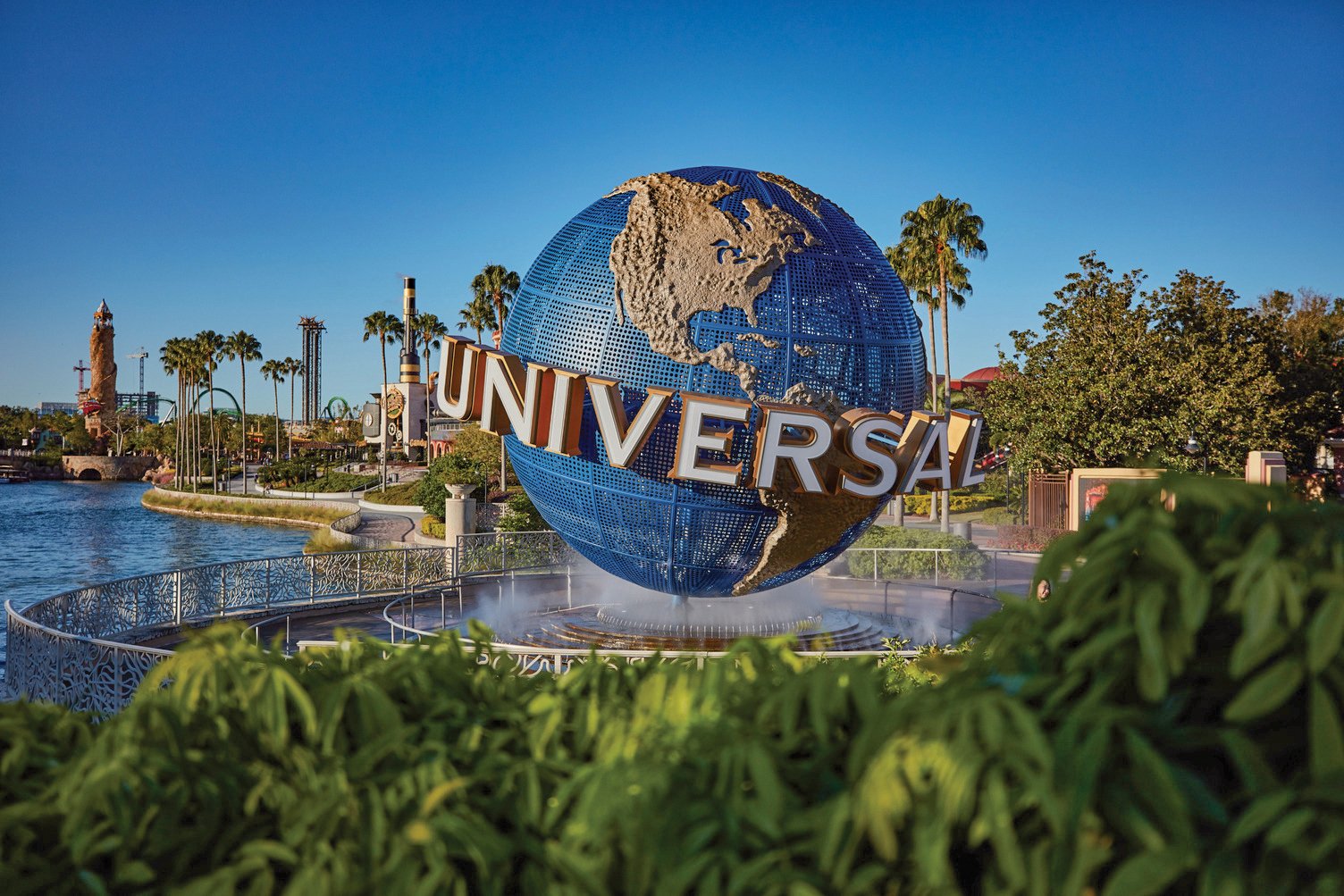 Universal Studios globe logo in a theme park setting with water, trees, and amusement park rides in the background.