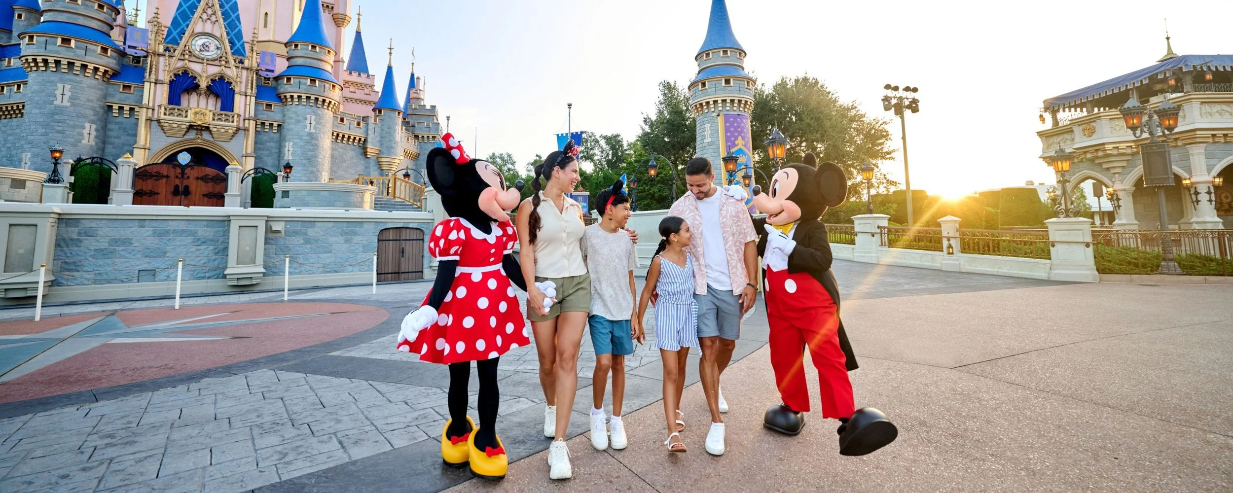 People, including children and adults, standing with Mickey Mouse and Minnie Mouse characters in front of Sleeping Beauty Castle at Disneyland during sunset.