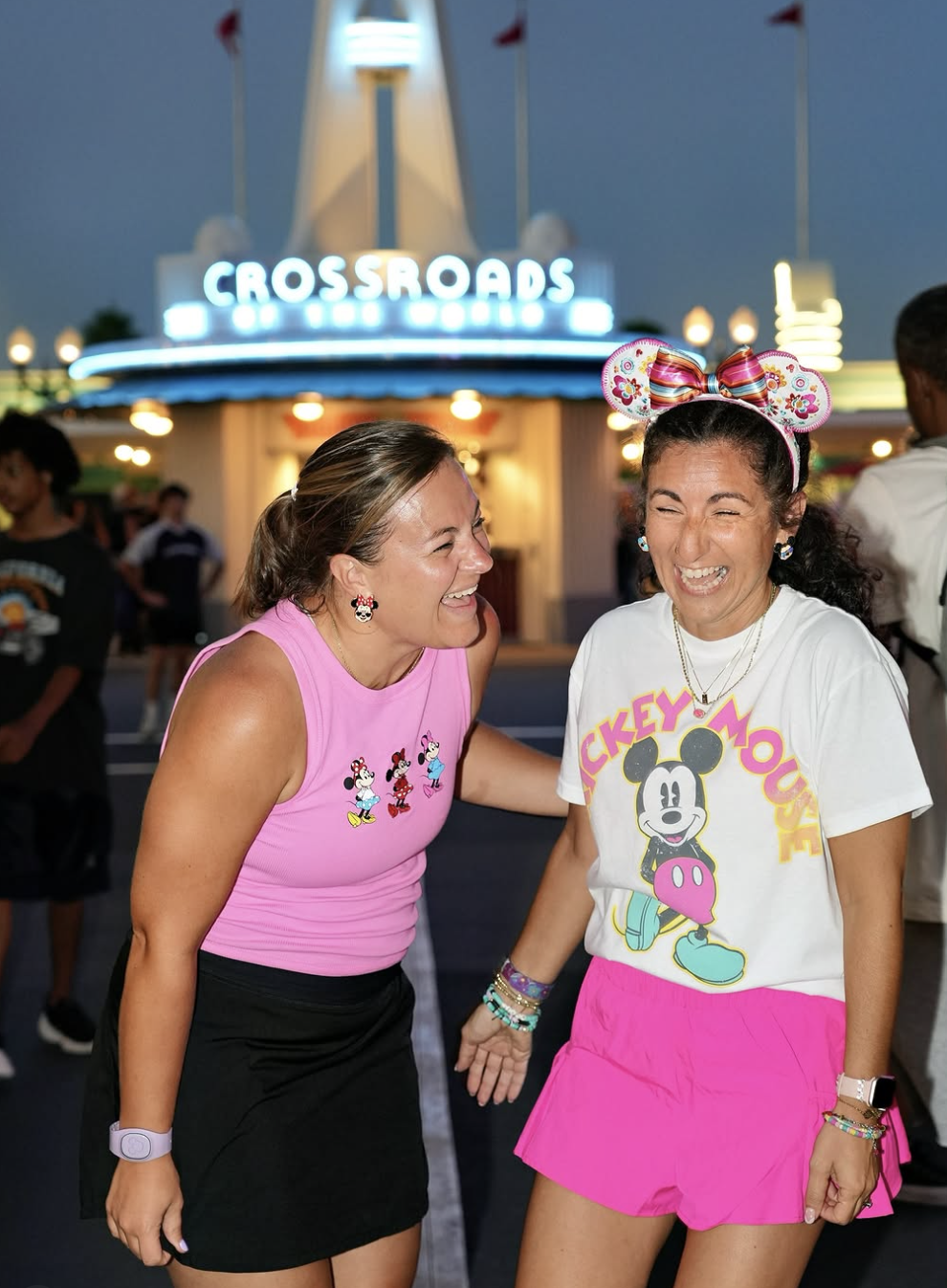 Two women laughing and having fun in front of an amusement park entrance at night. The woman on the left is wearing a pink sleeveless top with Minnie Mouse prints, a black skirt, and Minnie Mouse earrings. The woman on the right is wearing a white Mickey Mouse t-shirt, pink shorts, and Minnie Mouse ears headband. The background features bright lights and a sign that says 'CROSSLAND'.