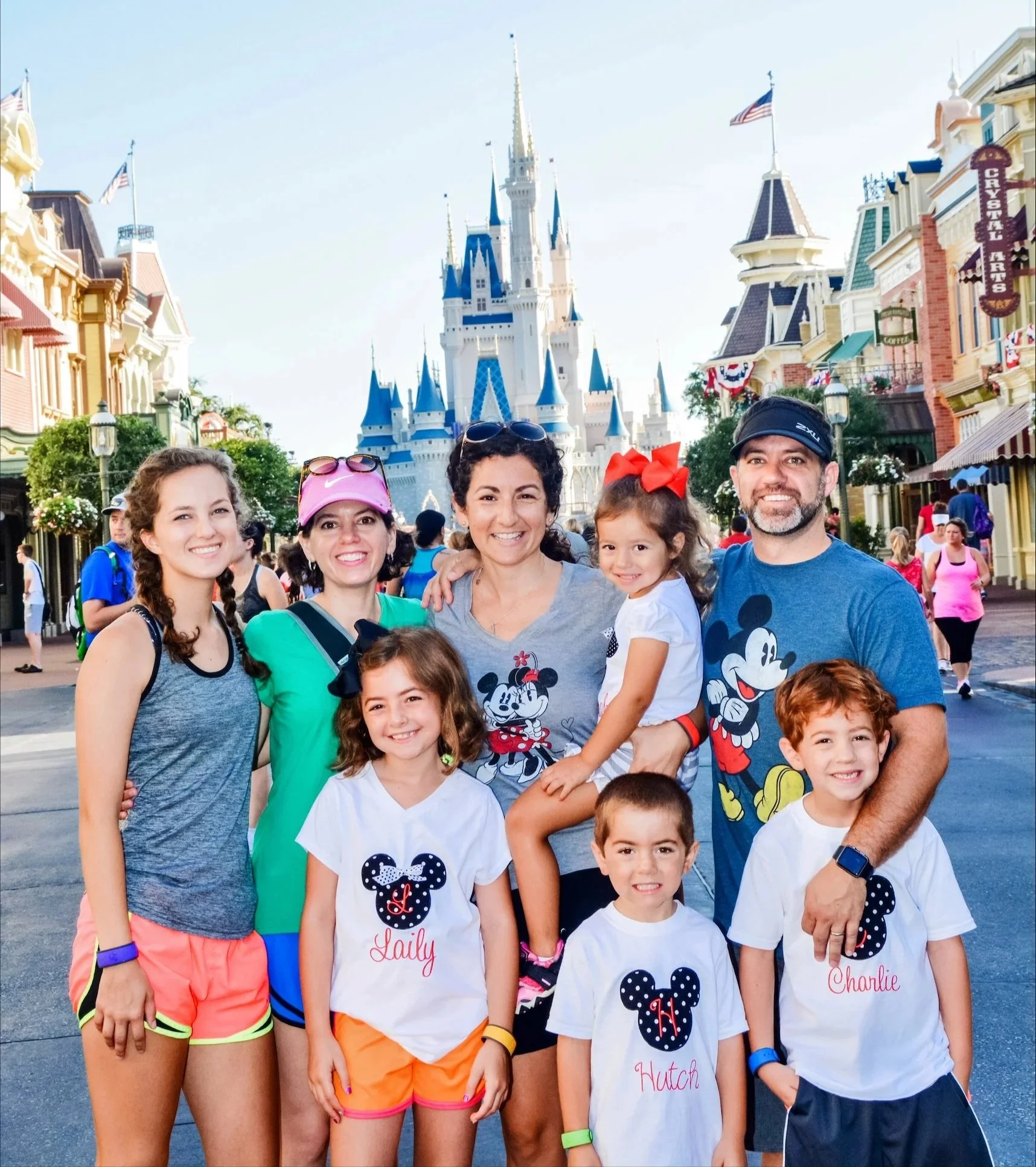 Group of five children and two adults standing in front of Sleeping Beauty Castle at Disneyland, with colorful buildings and American flags, some wearing Mickey Mouse themed T-shirts and accessories.