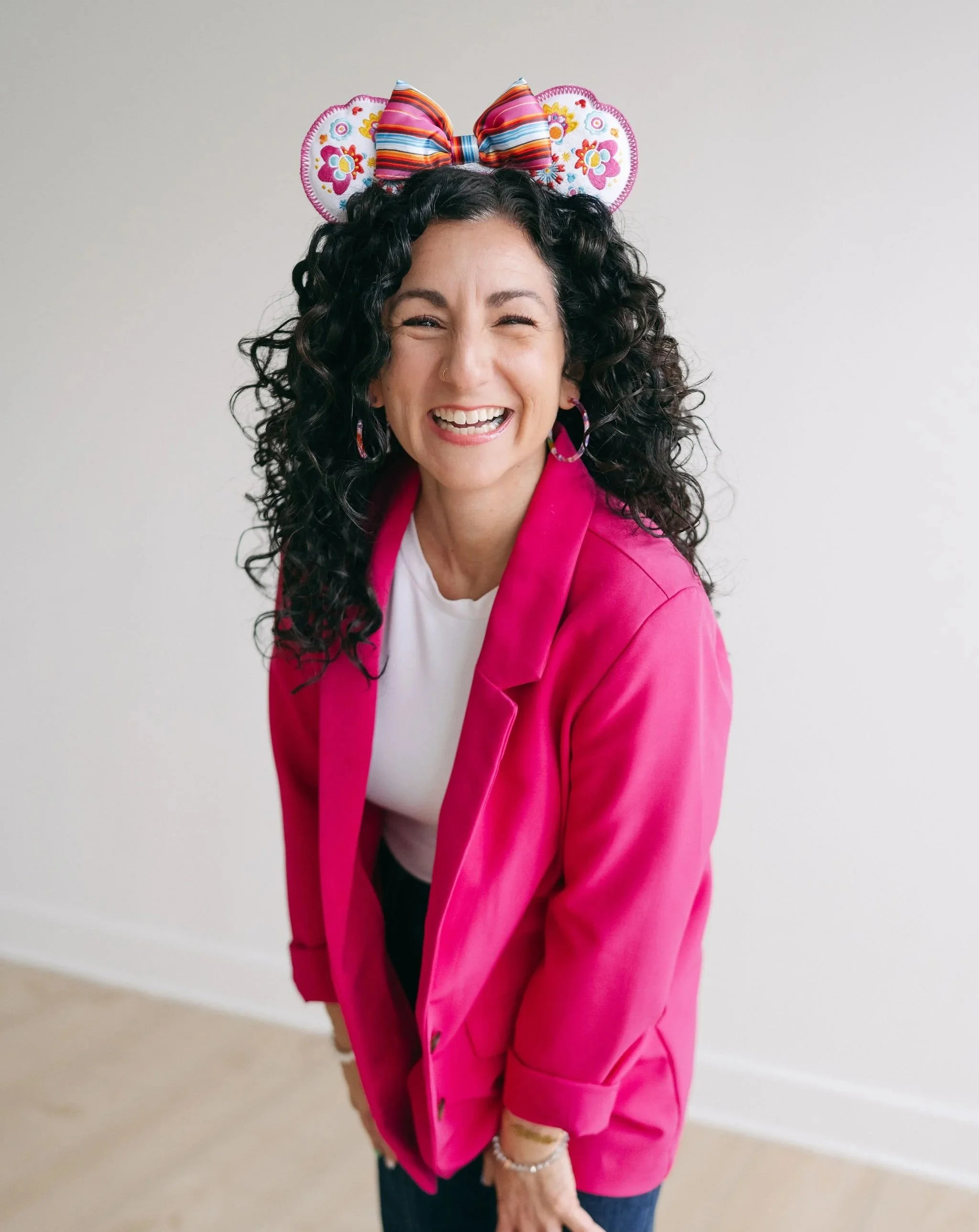 A woman with curly black hair smiling, wearing a pink blazer over a white t-shirt, with a colorful bow headband and earrings, standing against a neutral background.