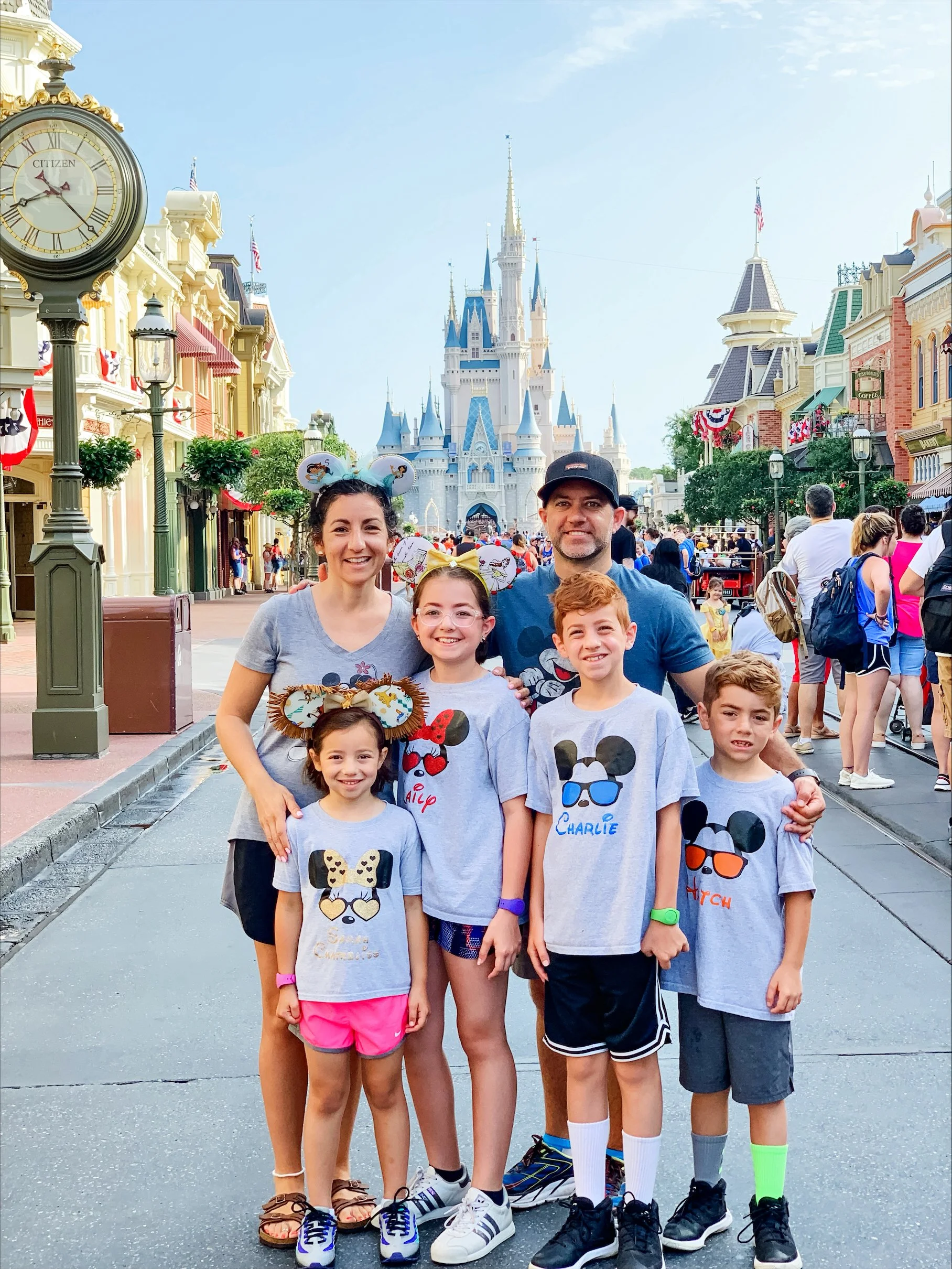 A family of seven standing in front of Sleeping Beauty Castle at Disneyland, wearing matching Mickey Mouse-themed T-shirts and Disney-themed accessories, with a crowd of visitors behind them and the castle in the background.