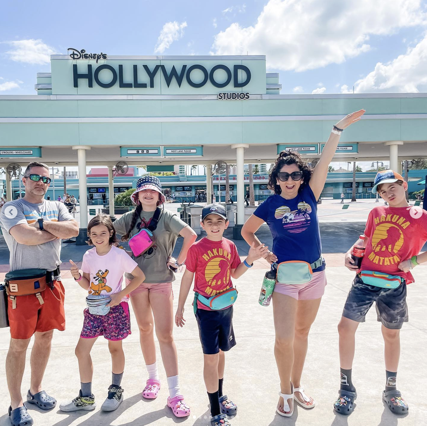 A group of six people, including children and adults, posing in front of Disney's Hollywood Studios entrance. They are smiling, wearing casual summer clothes, and standing on a sidewalk under a partly cloudy sky.