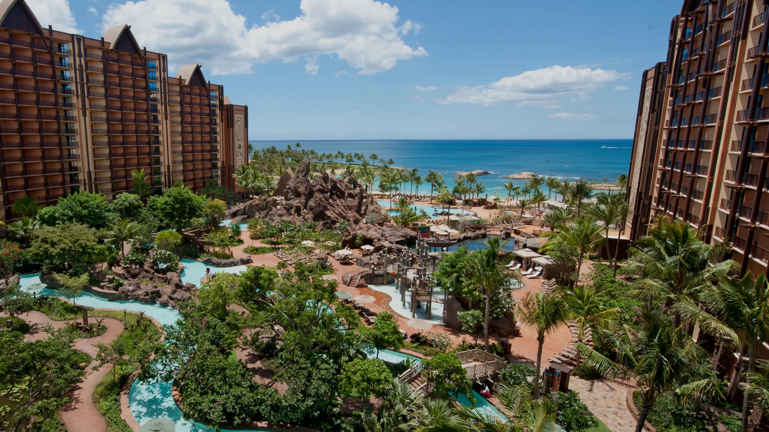 Scenic view of a tropical resort with lush greenery, a winding pool, and beachfront with the ocean in the background.