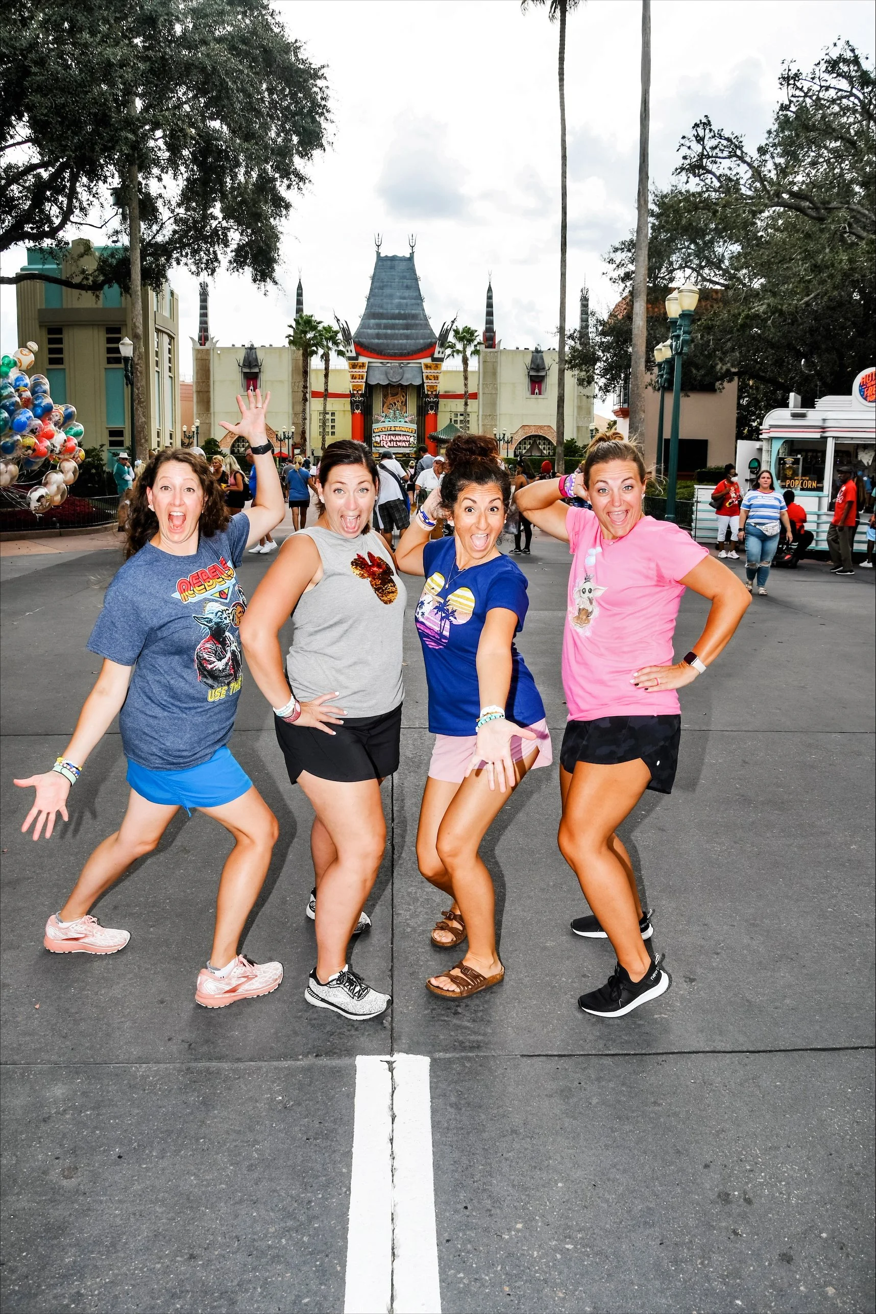 Four women standing on an outdoor street in front of a movie and theater themed attraction, striking playful poses. They are smiling and dressed casually in colorful t-shirts and shorts. Behind them there are trees, street lamps, and a crowd of people, with the Hollywood Tower Hotel attraction from Disney's Hollywood Studios in the background.