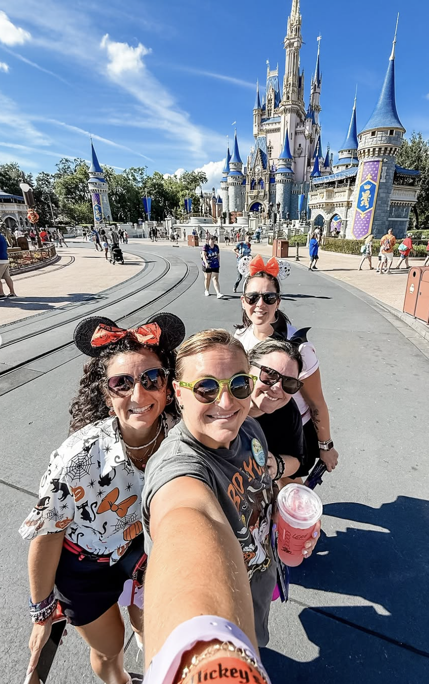 Group of four women taking a selfie in front of Cinderella's Castle at Disney theme park, wearing Mickey Mouse ears and sunglasses, on a sunny day.