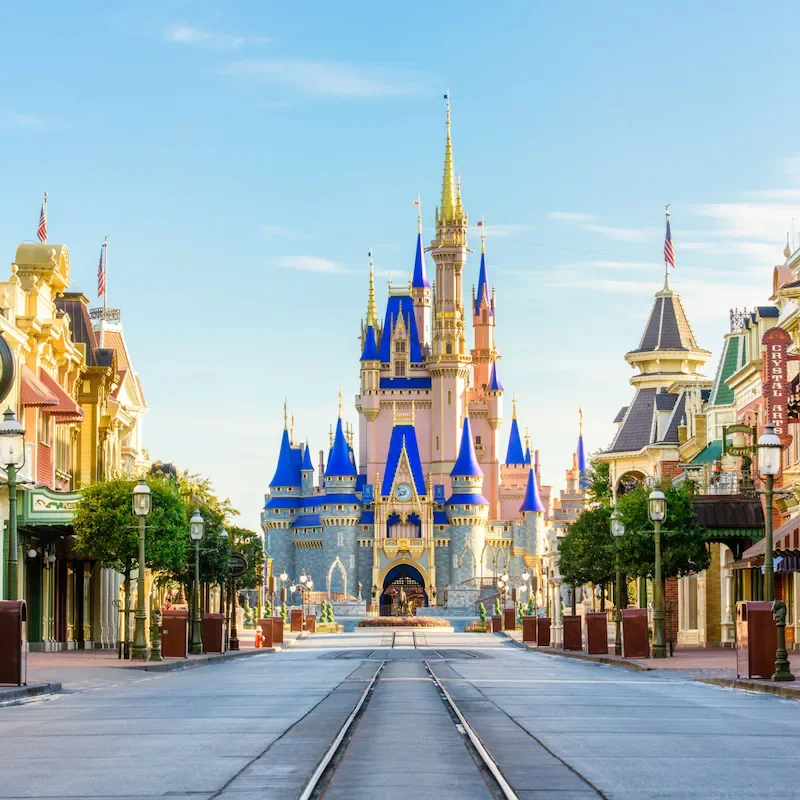 Empty Main Street with Sleeping Beauty Castle at Disneyland in the distance, clear blue sky, colorful buildings, and tram tracks in the foreground.