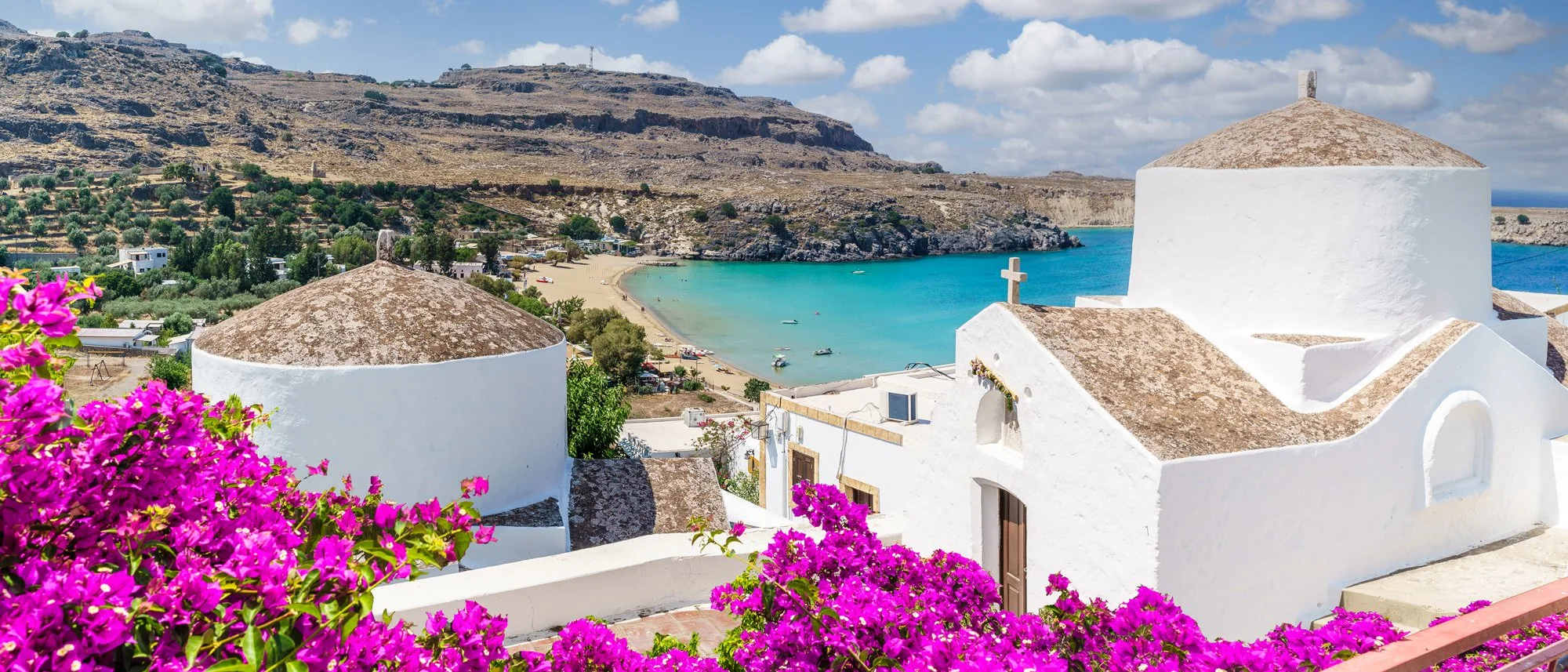 A scenic view of a Greek island with white churches, vibrant pink flowers in the foreground, a sandy beach, turquoise sea, and rocky hills in the background under a partly cloudy sky.
