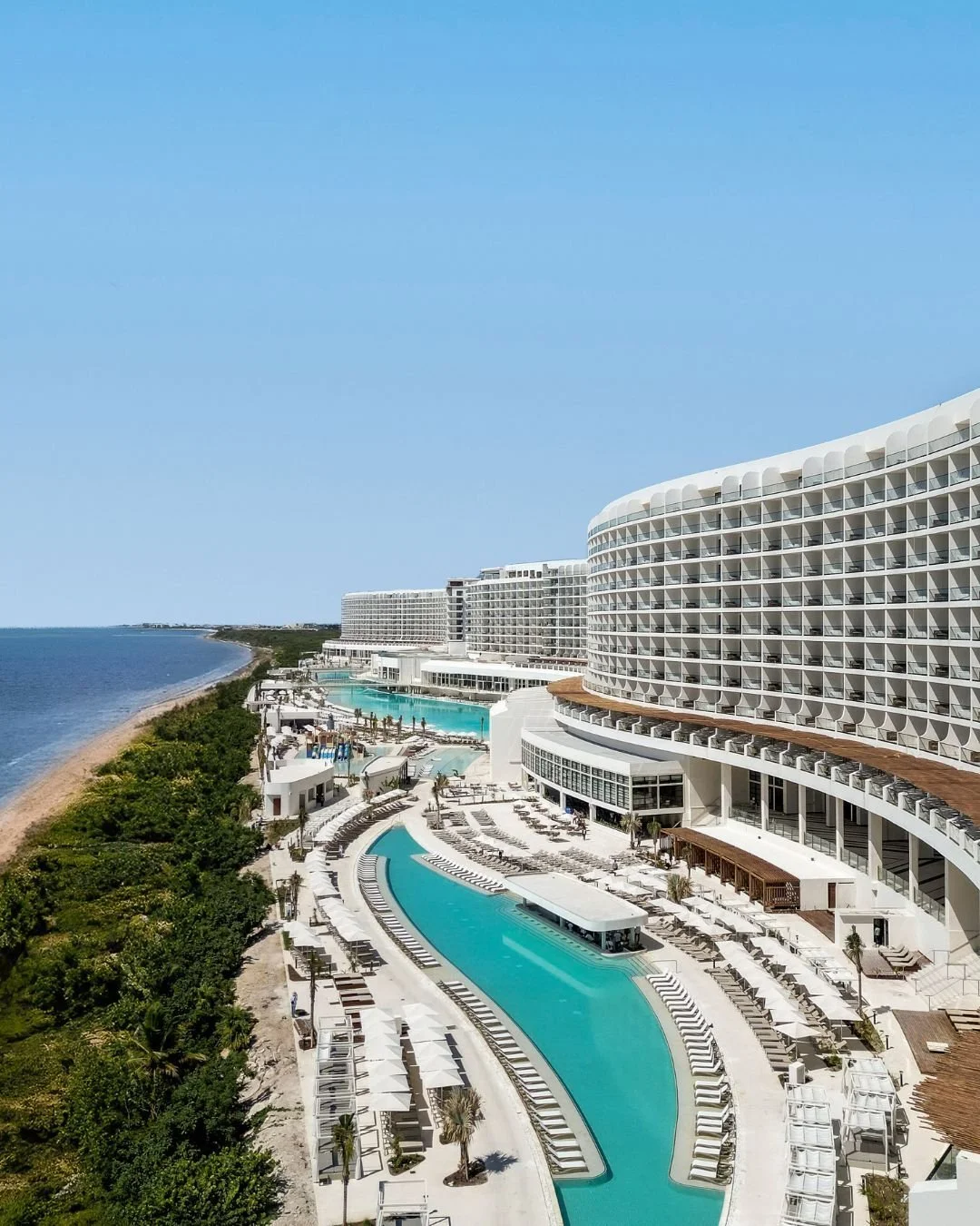 Luxury beachfront resort with multiple white high-rise buildings, outdoor pools, sun loungers, and a curved swimming pool near the ocean under a clear blue sky.
