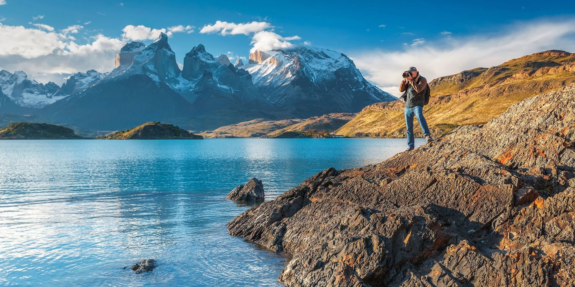 A person standing on rocky shoreline taking a photograph of snow-covered mountains and a lake in the background.