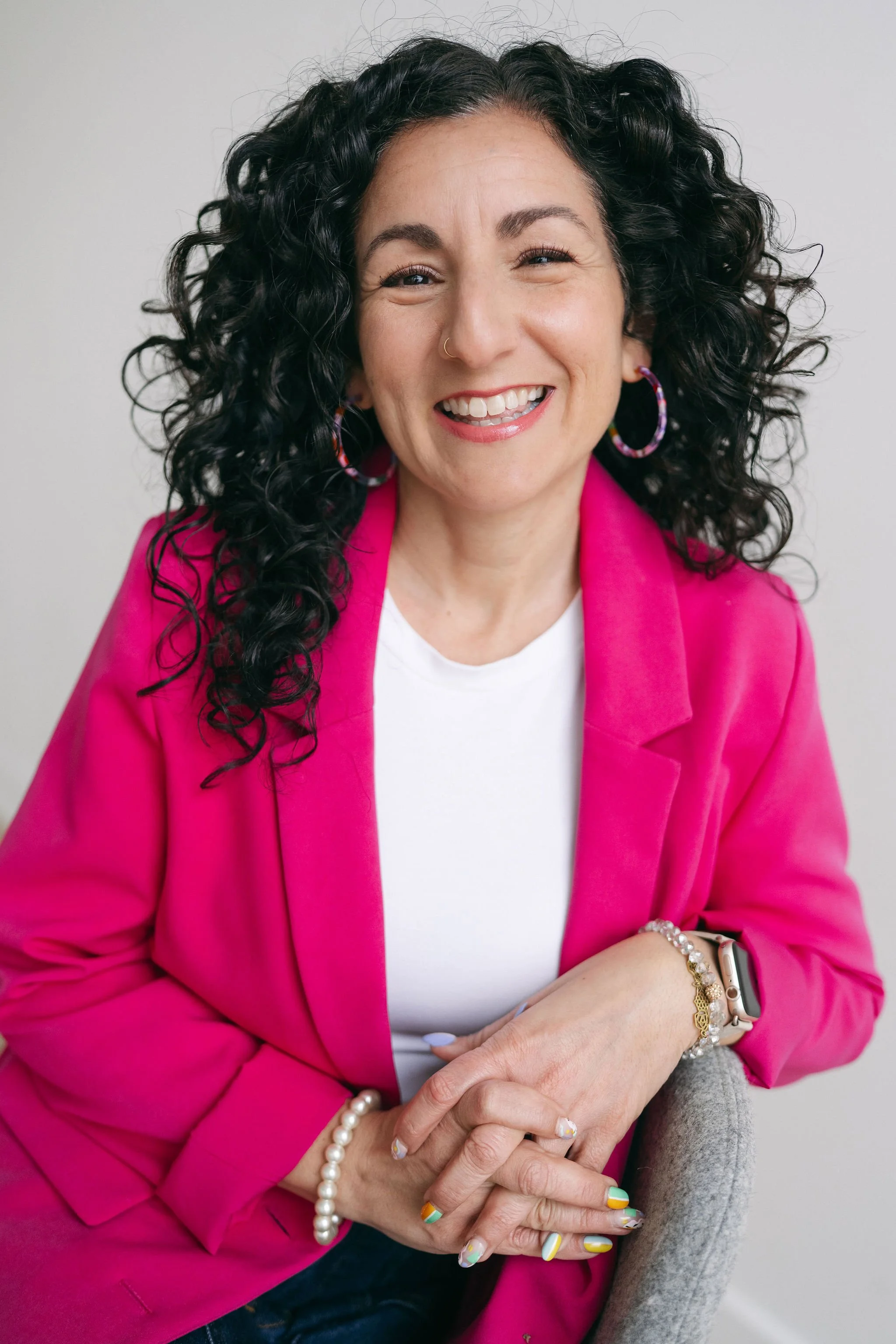 A woman with curly black hair, wearing a pink blazer, white top, colorful hoop earrings, and various bracelets and rings, sitting on a gray chair, smiling.