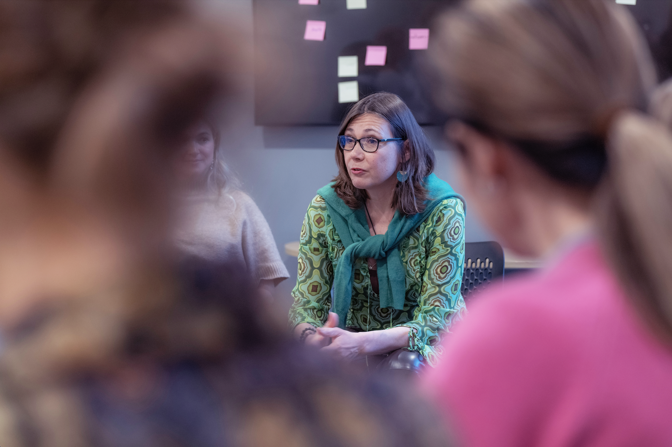 A woman with glasses and shoulder-length brown hair, wearing a green patterned shirt and teal scarf, is sitting and speaking in a group setting, with a blackboard with sticky notes in the background. The image is framed by blurred people in the foreground.