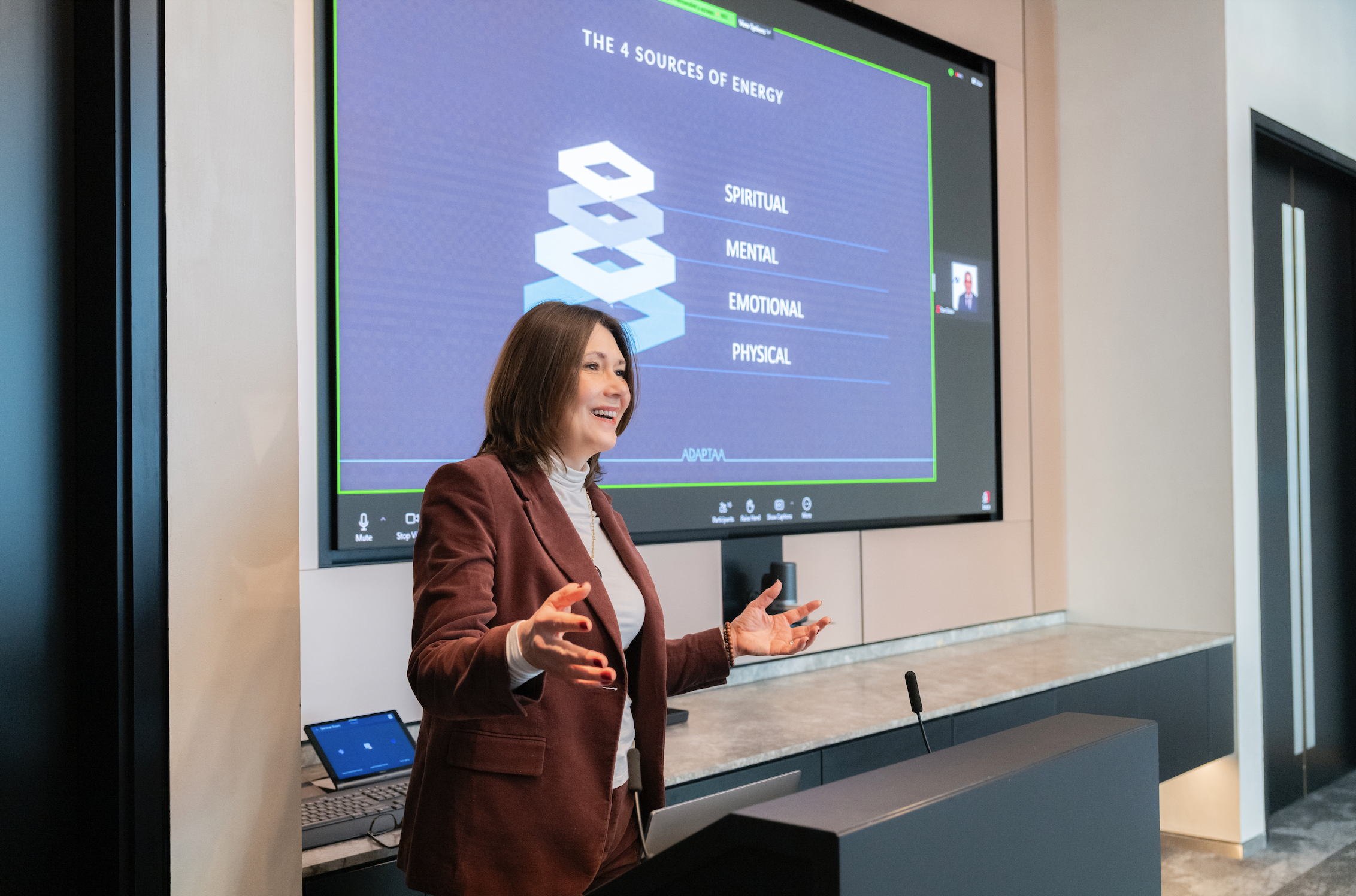 A woman giving a presentation in front of a screen displaying a slide titled 'The 4 Sources of Energy,' showing a diagram with four levels labeled 'Spiritual,' 'Mental,' 'Emotional,' and 'Physical.' She is smiling and gesturing with her hands.