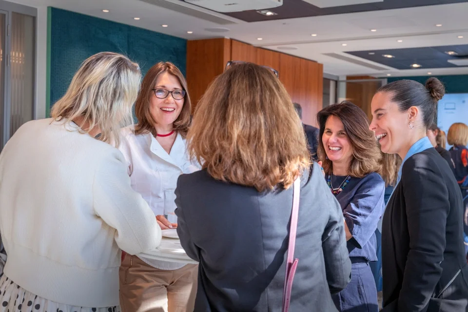 A group of five women in a professional setting engaged in a lively conversation, smiling and laughing.